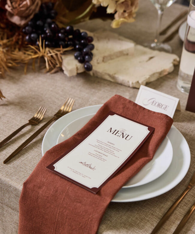 Elegant table setting with a white plate, a rust-colored cloth napkin, a menu card, and gold utensils.