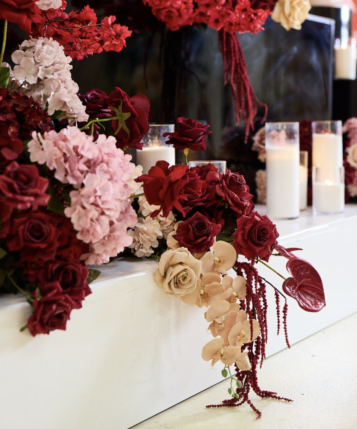 Arranged floral display with red, pink, cream roses, orchids, and other flowers, decorated with candles in glass holders on a white surface.