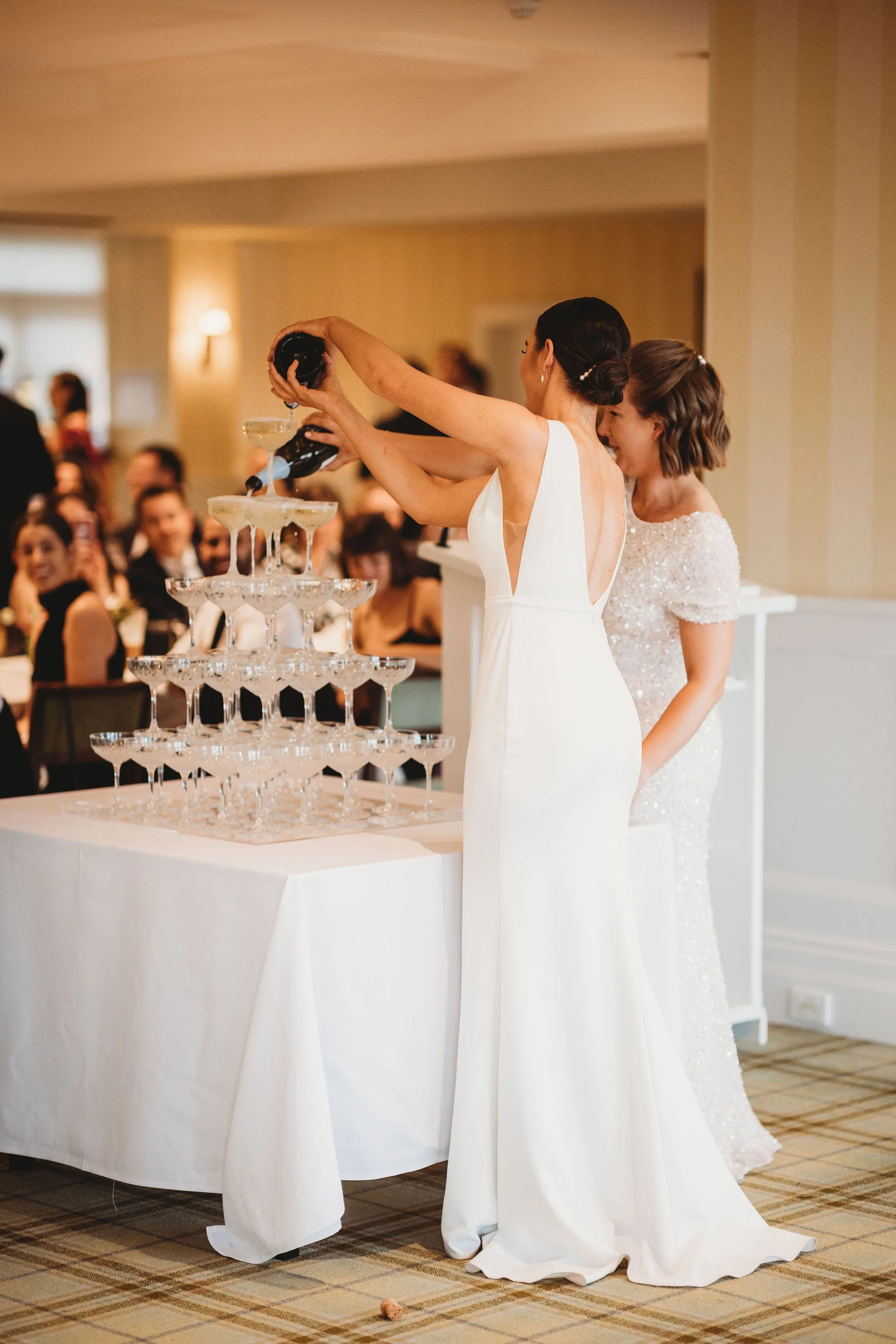 A bride in a white wedding gown pouring champagne into a tower of glasses at a wedding reception, with guests watching and smiling in the background.
