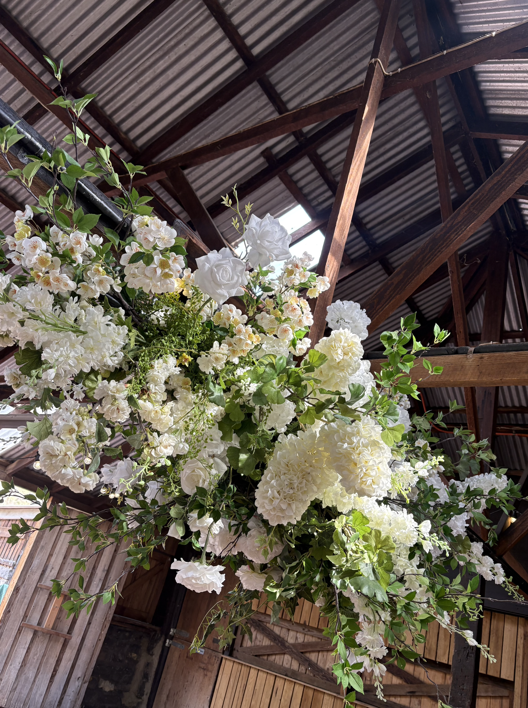 A hanging arrangement of white flowers and green leaves inside a rustic barn with a wooden ceiling and metal roof.