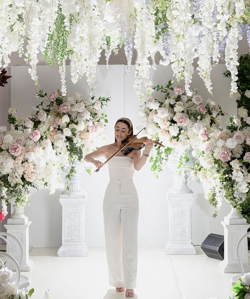 A woman in white pants and a strapless top playing a violin in a flower-decorated setting with hanging white flowers and large floral arrangements.