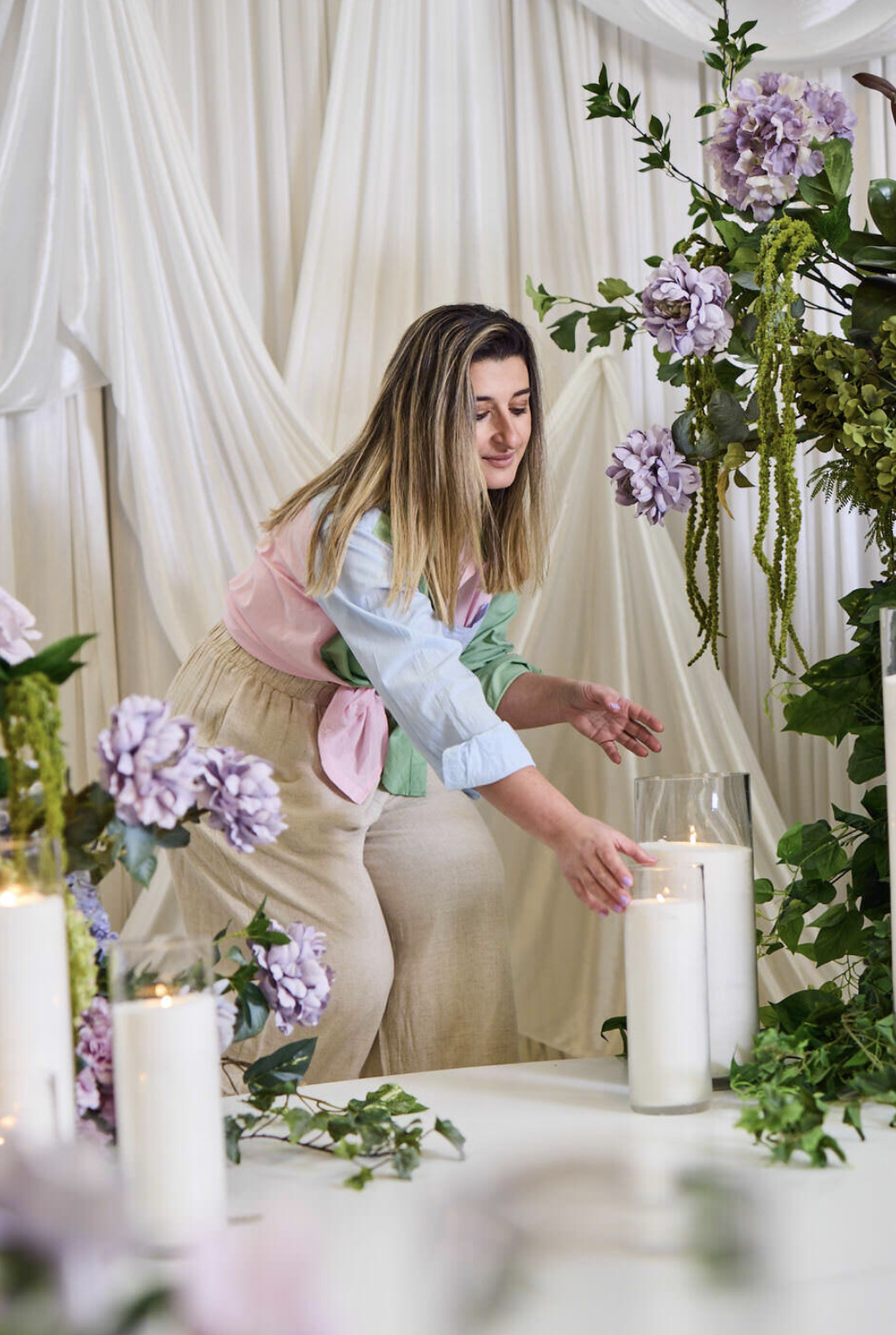 Woman arranging large white candles with flowers and greenery in a decorated setting.