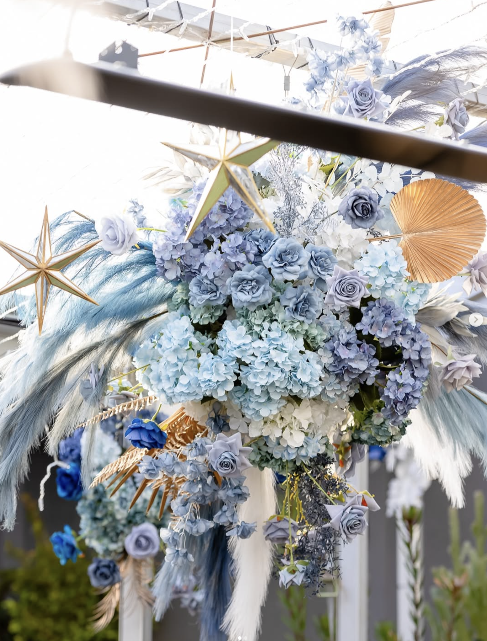 Decorative floral arrangement with blue, white, and purple flowers, gold leaves, and hanging gold star and fan ornaments.