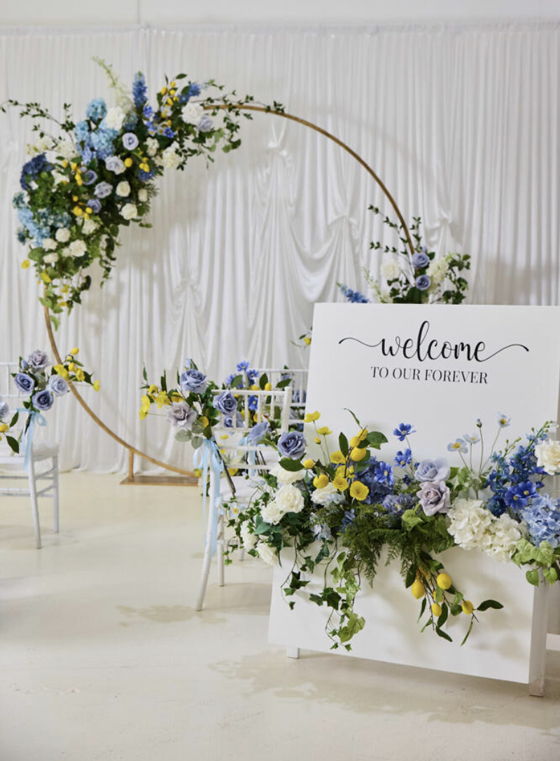 A man standing on a step ladder arranging a large floral display with white, blue, and yellow flowers against a white pleated backdrop.