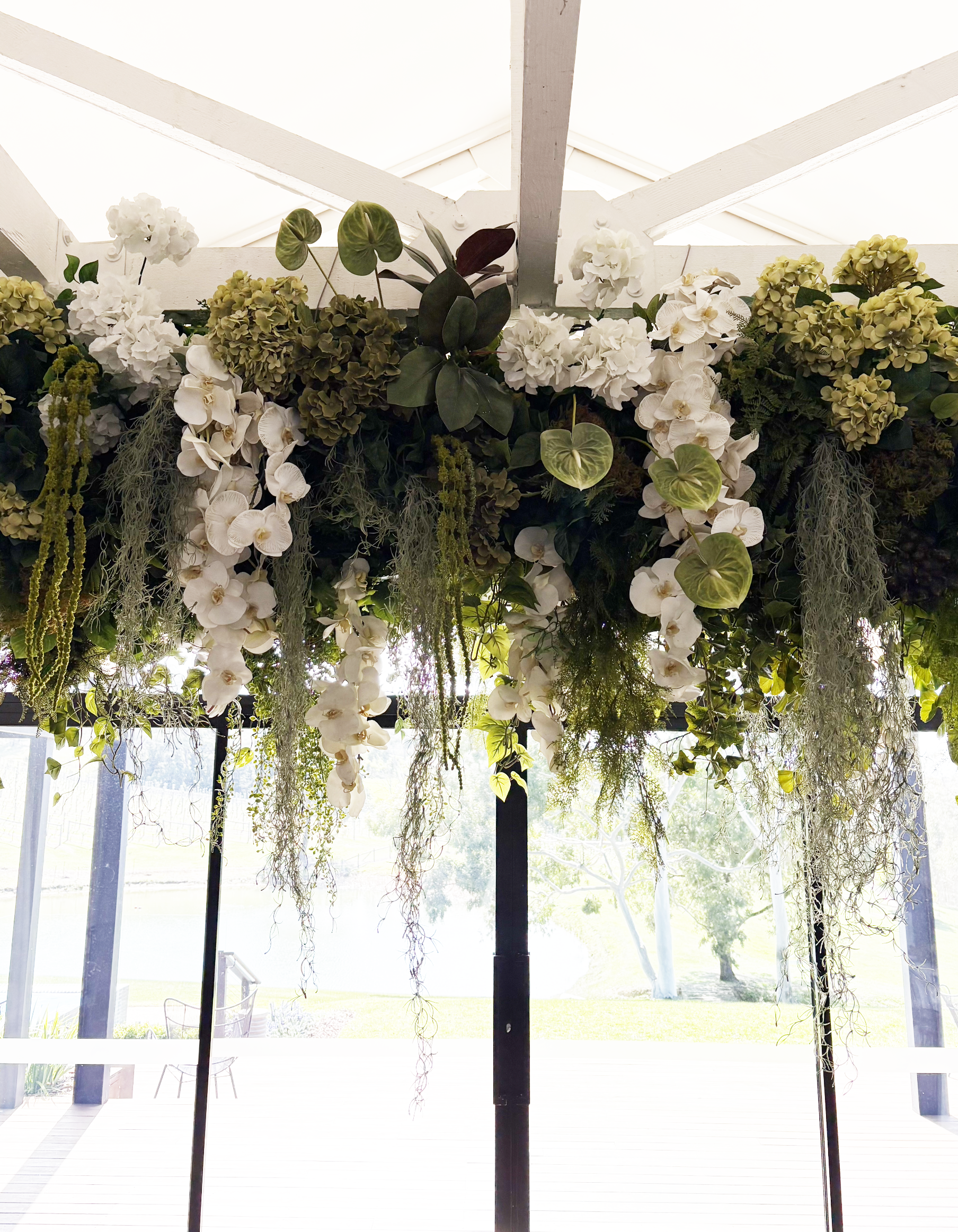 Overhead floral arrangement with white hydrangeas, orchids, and various greenery hanging from a white ceiling at an outdoor venue.