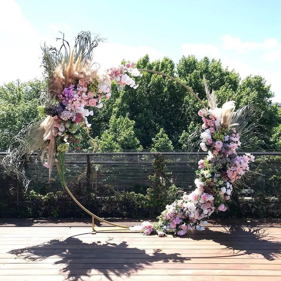 A floral arch with pink, white, and purple flowers, and dried pampas grass, set outdoors on a wooden deck with trees and a fence in the background.