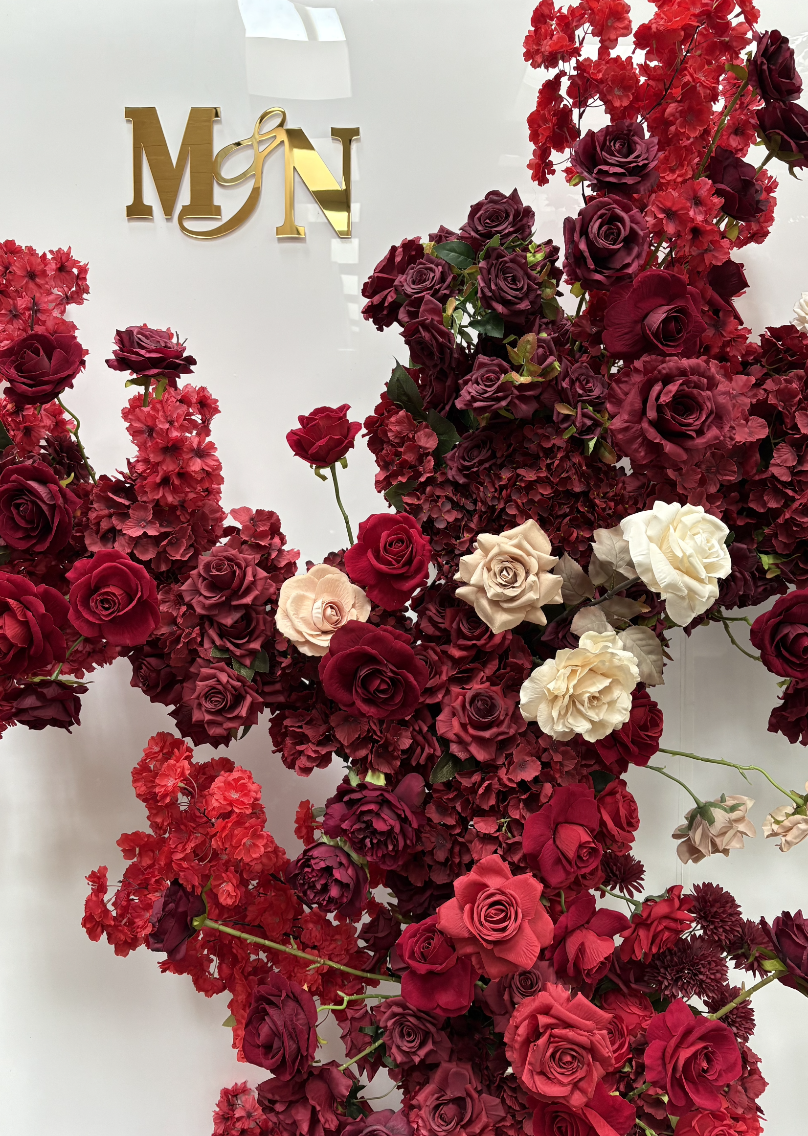 A display of various red, pink, purple, and white roses with greenery, arranged around a white background with a gold sign that reads 'MGN' in the upper left corner.