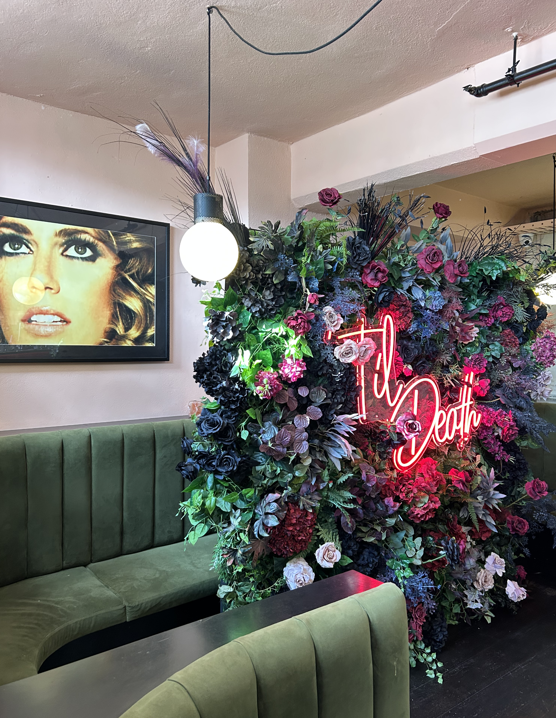 A colorful floral wall with a neon sign that reads 'F**k Death,' situated next to a green upholstered booth and a wall-mounted framed photo of a woman.