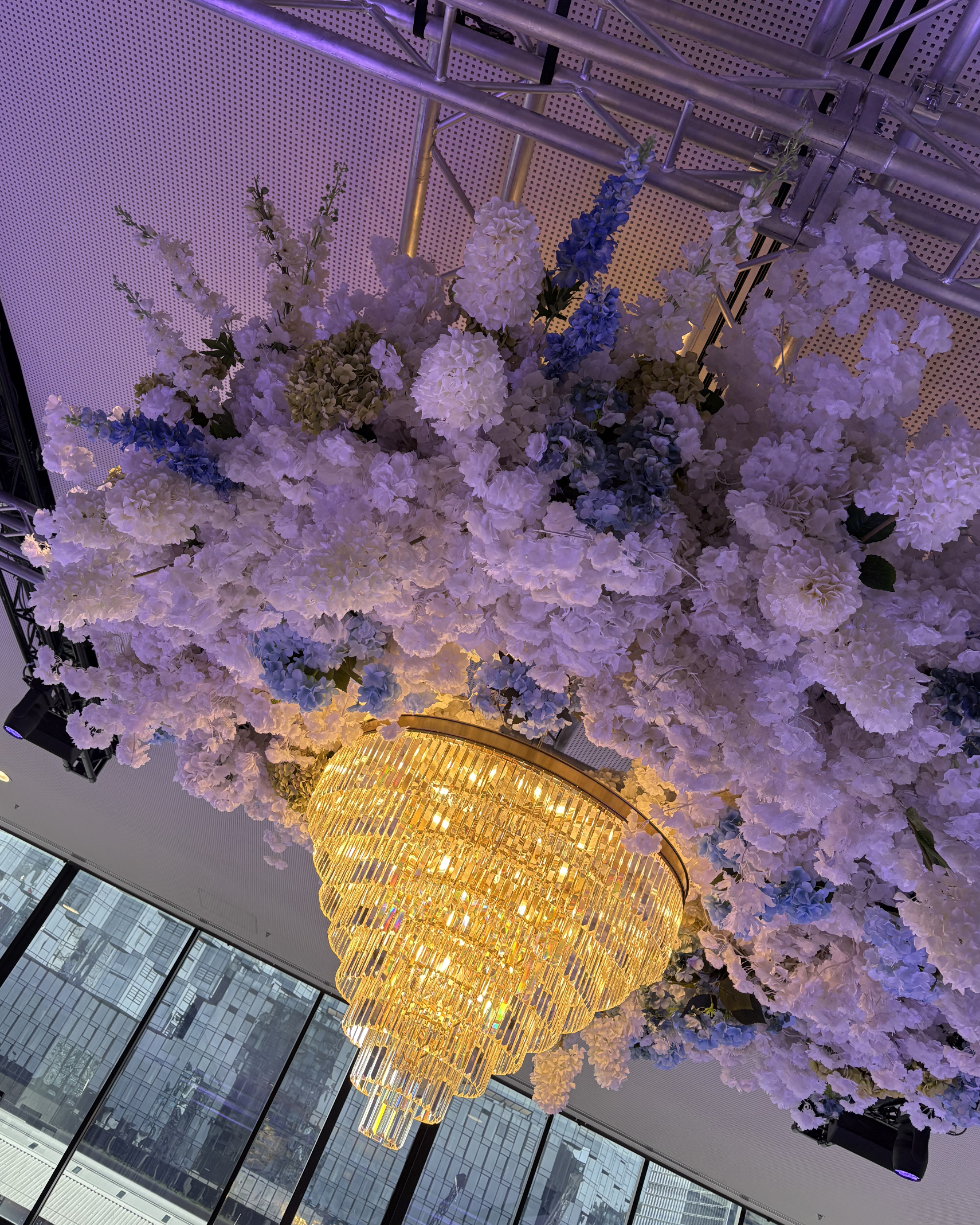 A large, ornate gold chandelier hanging from the ceiling surrounded by an arrangement of white, purple, and blue flowers.