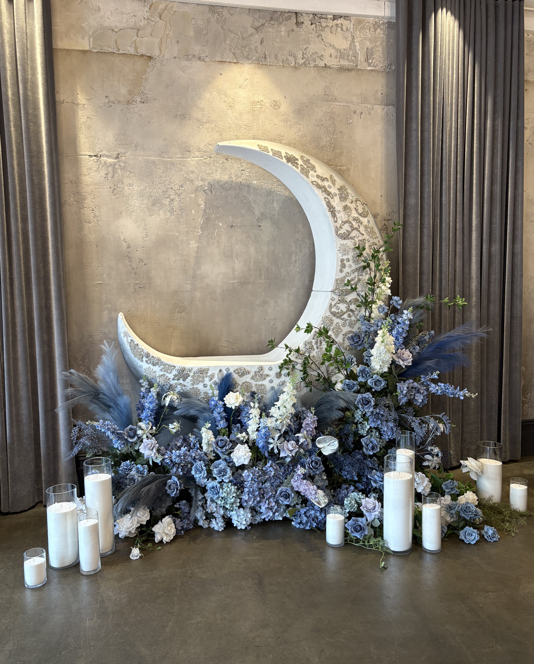 Decorative moon-shaped art piece with floral arrangement in shades of blue, white, and gray, surrounded by white pillar candles on the floor.