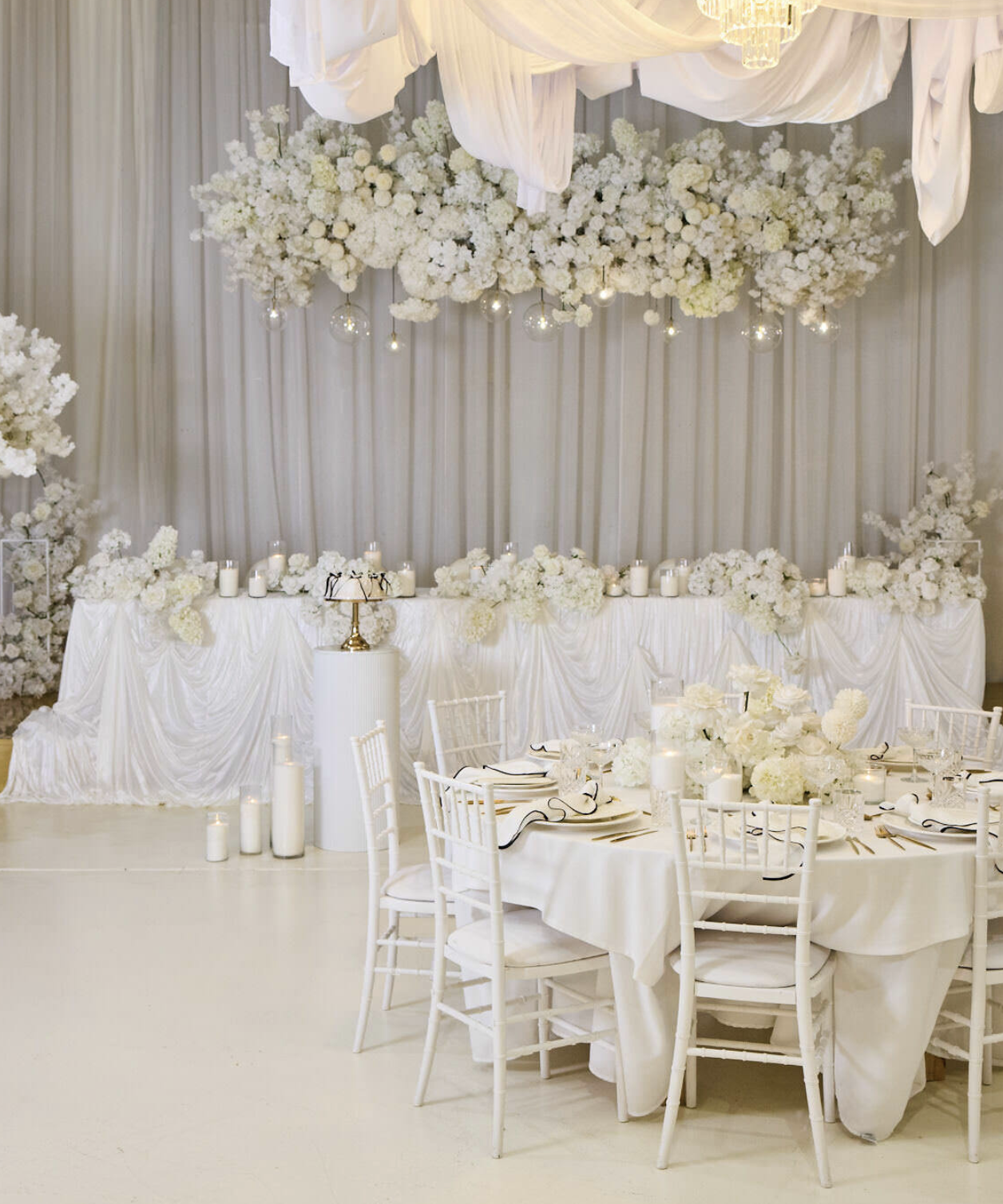 Elegant wedding reception setup with a long head table decorated with white flowers, candles, and drapery backdrop, and a round table with white chairs and floral centerpieces.