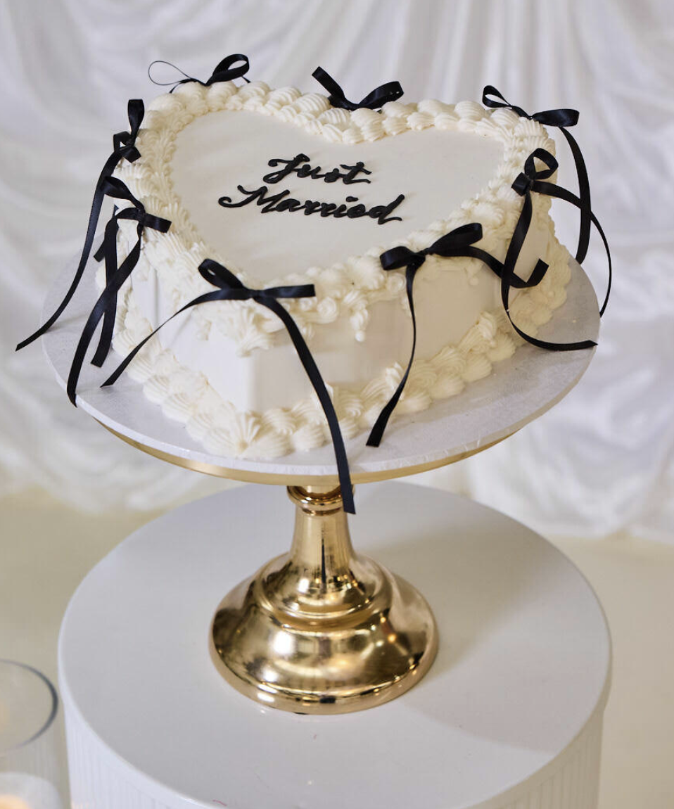 Heart-shaped wedding cake with white frosting, black ribbon decorations, and the words "Just Married" written on it, displayed on a gold cake stand.