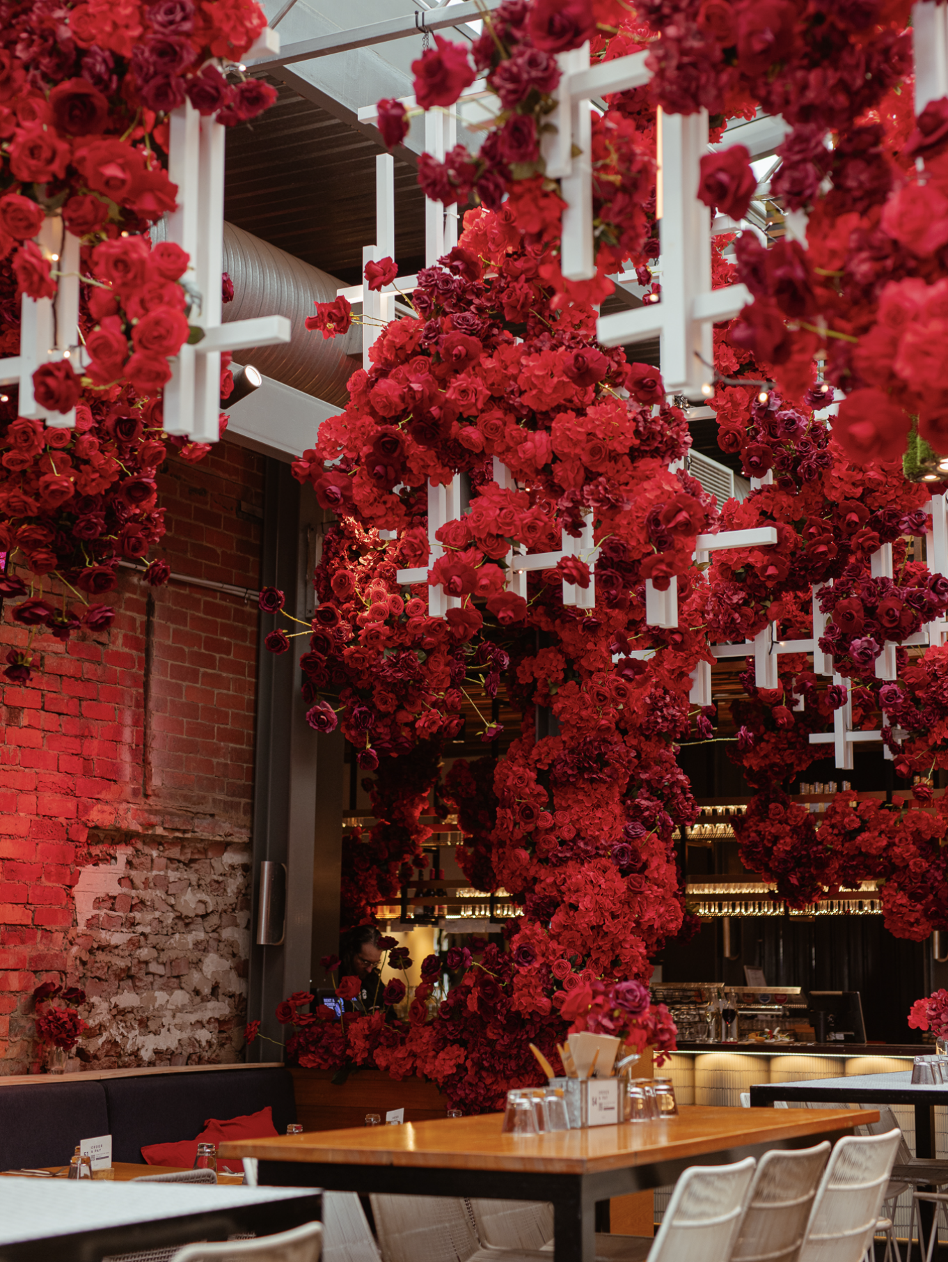 Interior of a restaurant decorated with abundant red floral arrangements hanging from white framework, exposing exposed brick walls and modern furnishings.