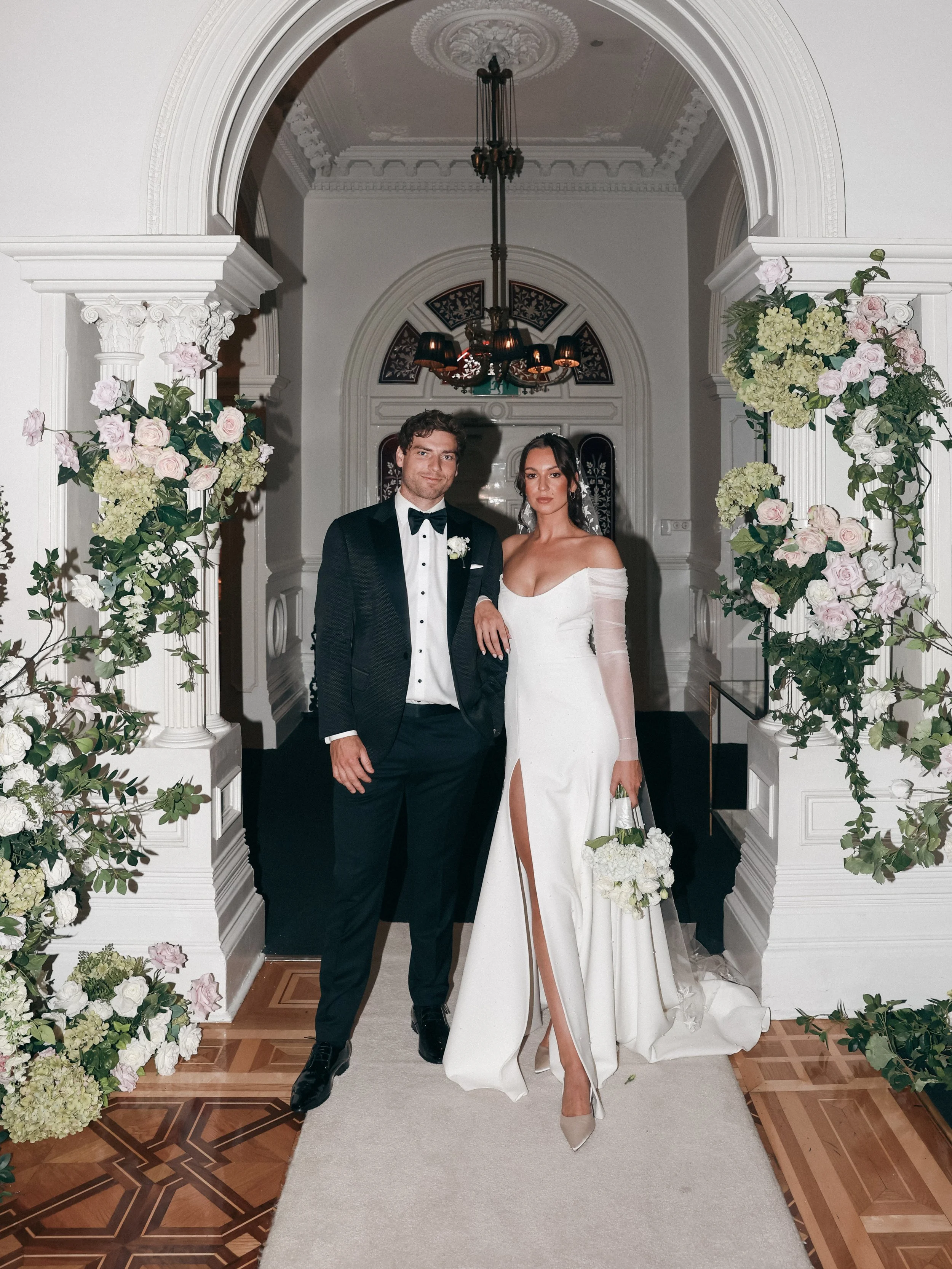 A bride and groom on their wedding day standing under a floral archway in a luxurious interior with a chandelier.