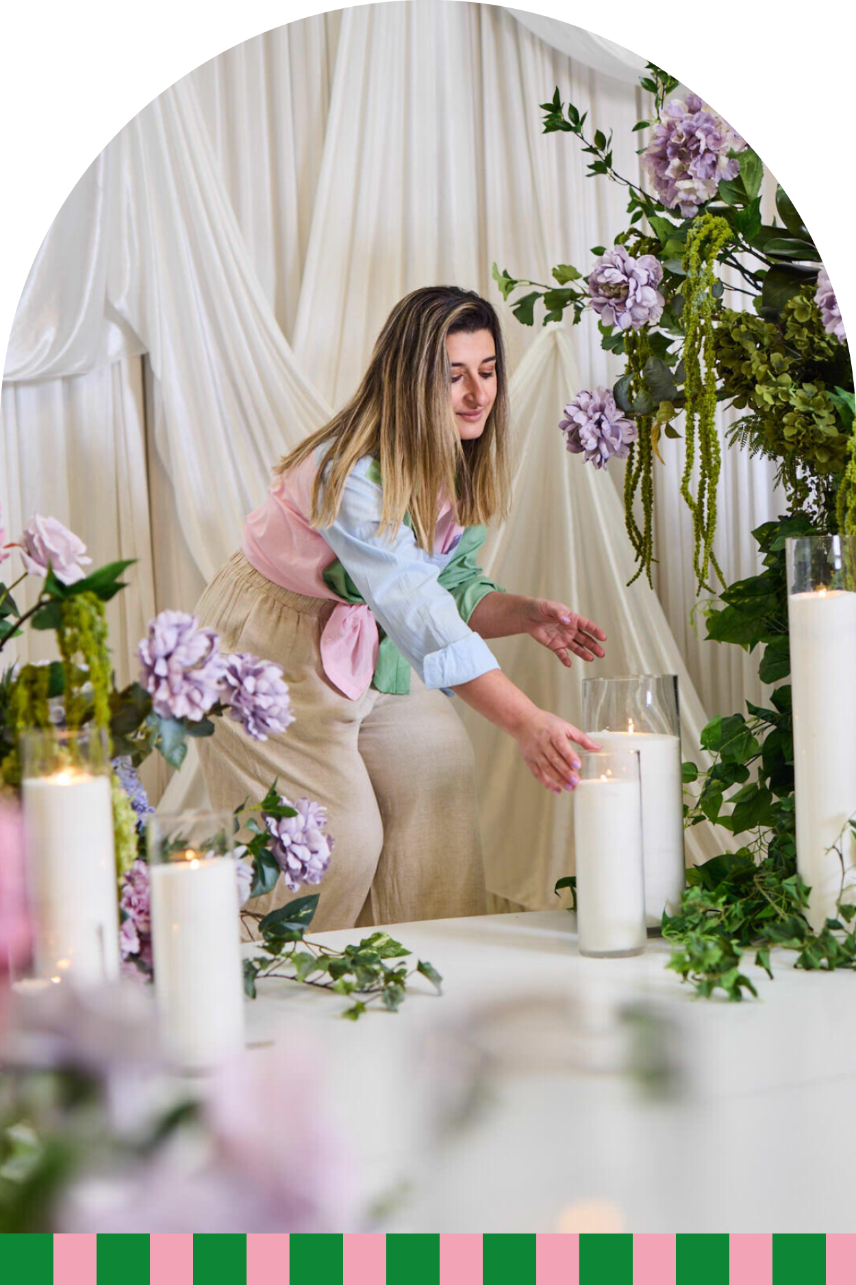 A woman arranging white candles surrounded by purple flowers and green foliage on a table with a cream-colored draped curtain backdrop.