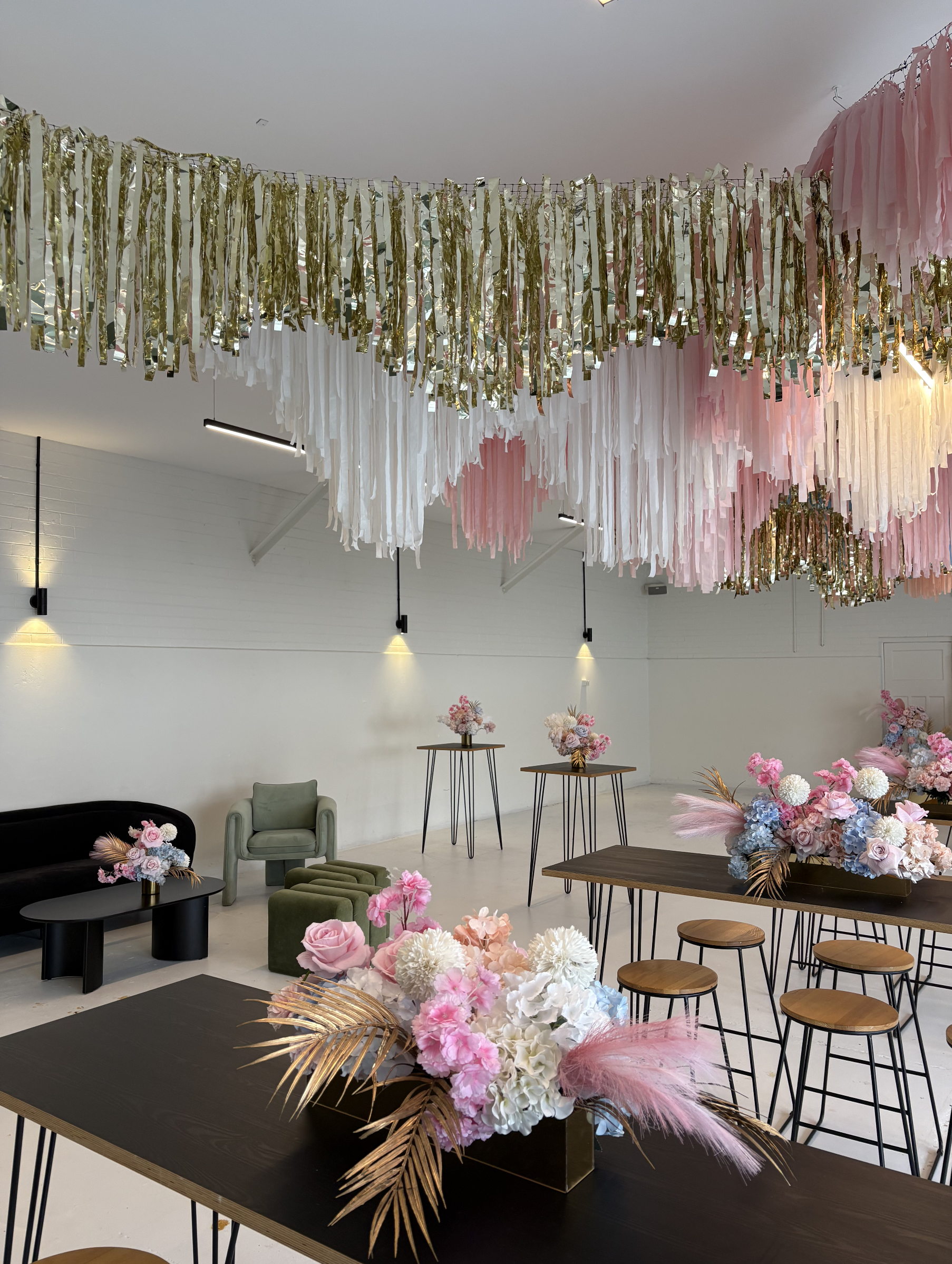 Decorated event space with pink, white, and gold streamers hanging from the ceiling, floral arrangements with pink, white, and blue flowers on tables and a sofa, and minimal lighting.