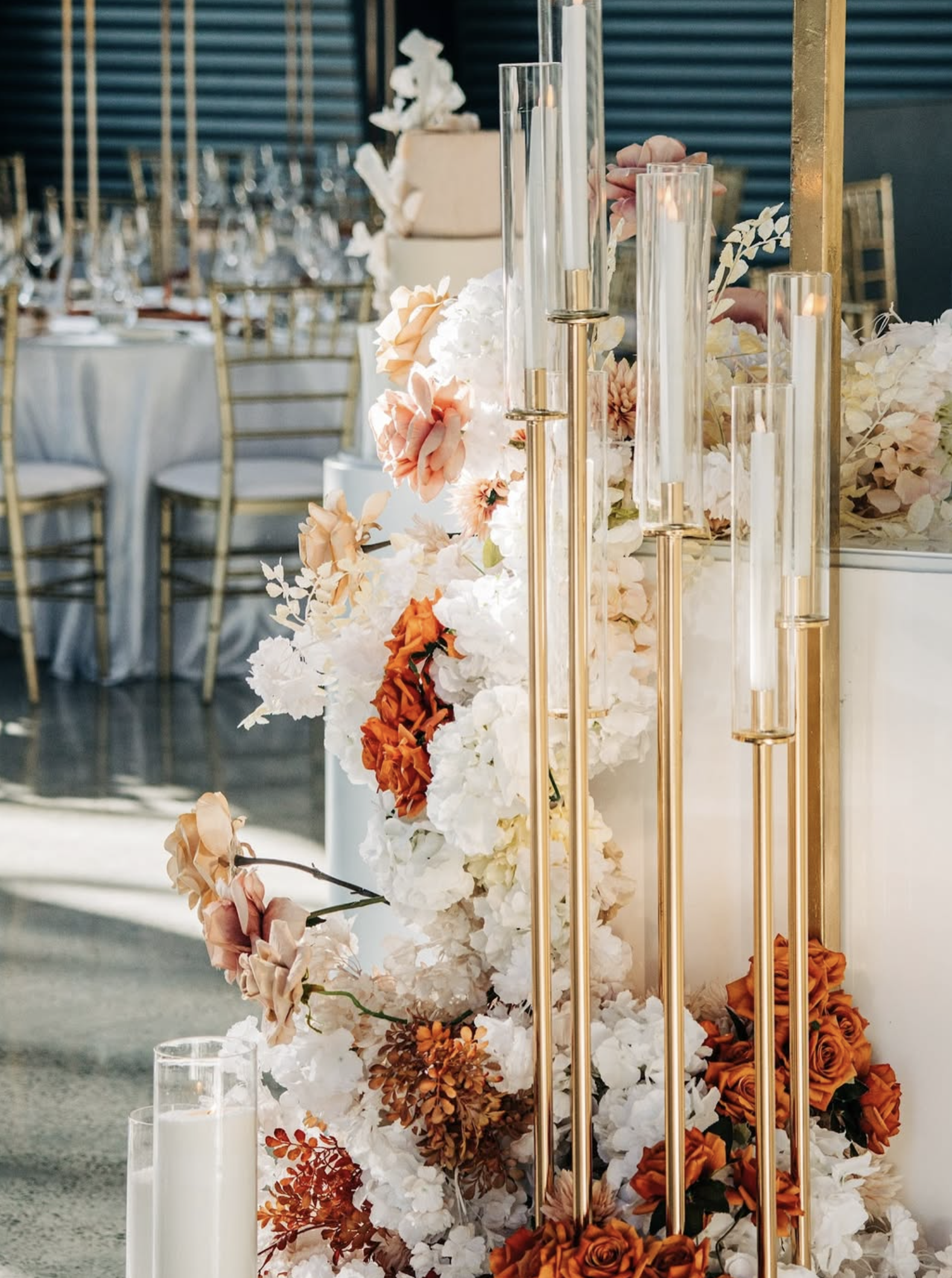 Elegant floral wedding decor with white, blush, and orange flowers, tall glass candle holders with lit white candles, and gold accents in a reception hall.