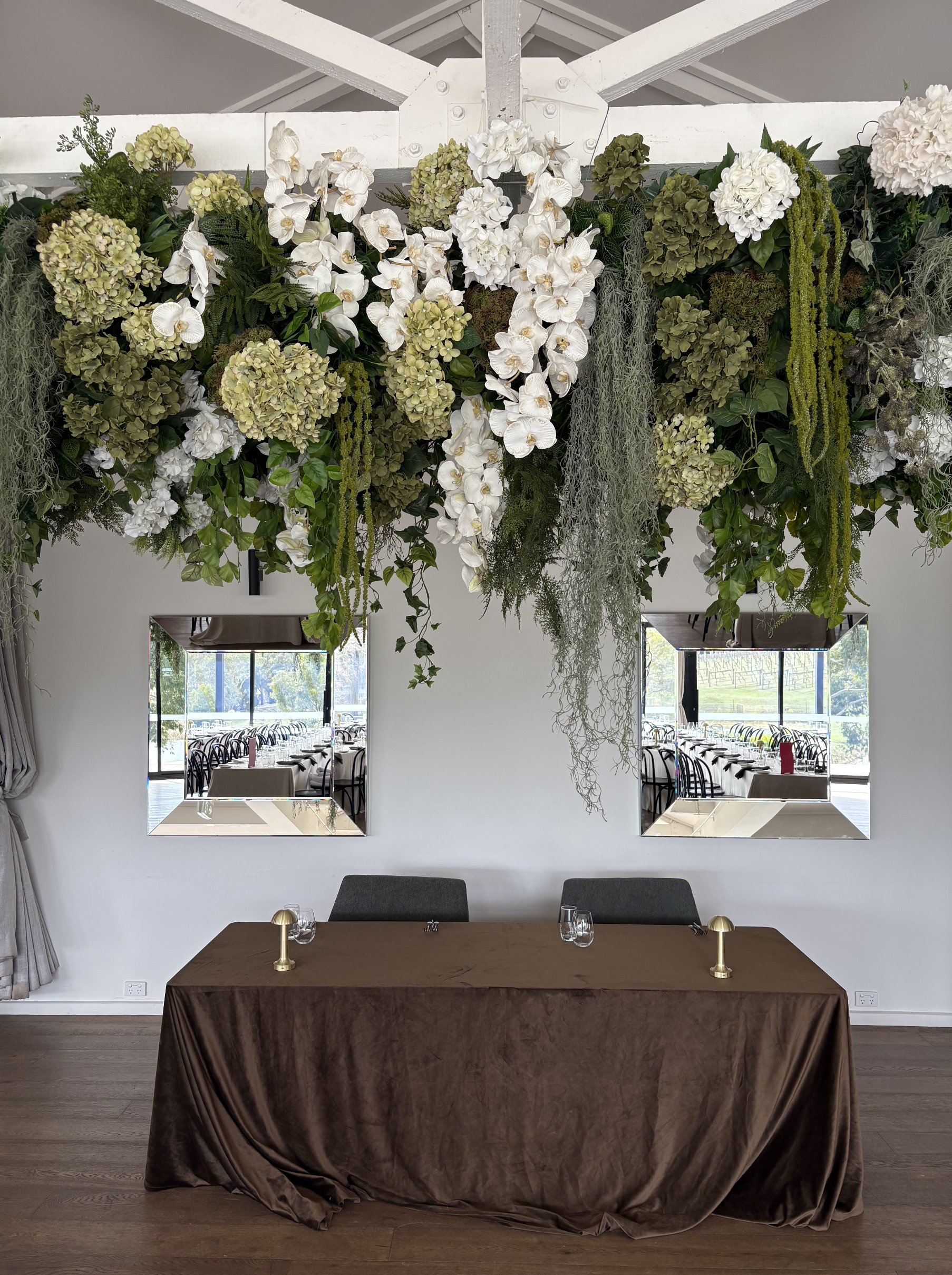 Interior of a decorated dining area with a large floral arrangement hanging from the ceiling, featuring white and green flowers. There are two windows behind a table with a brown tablecloth, set with glasses and brass lamps.