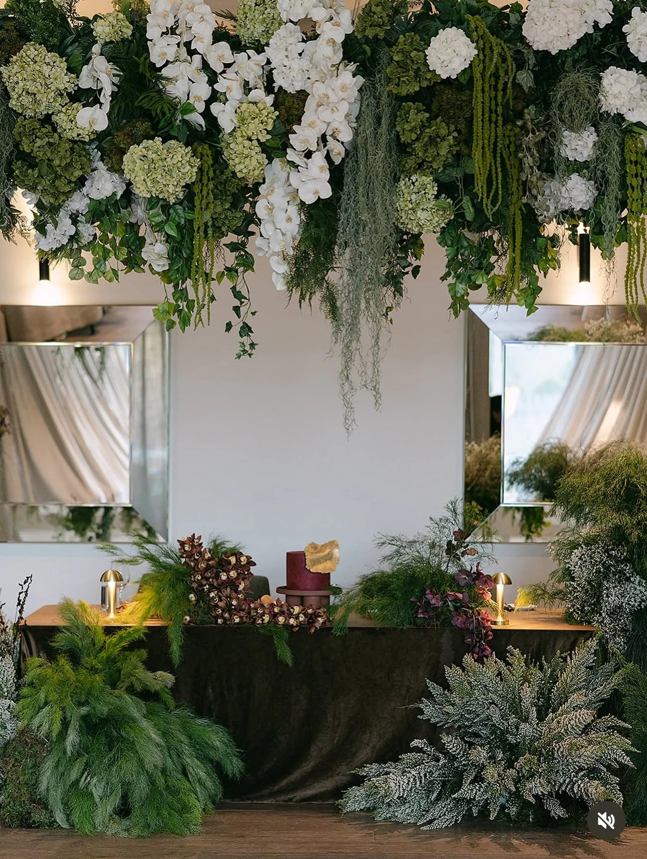 Elegant event setup with floral arrangements including white orchids, hydrangeas, and greenery, reflected in two large mirrors, with a table decorated with a burgundy cake, candles, and lush foliage.