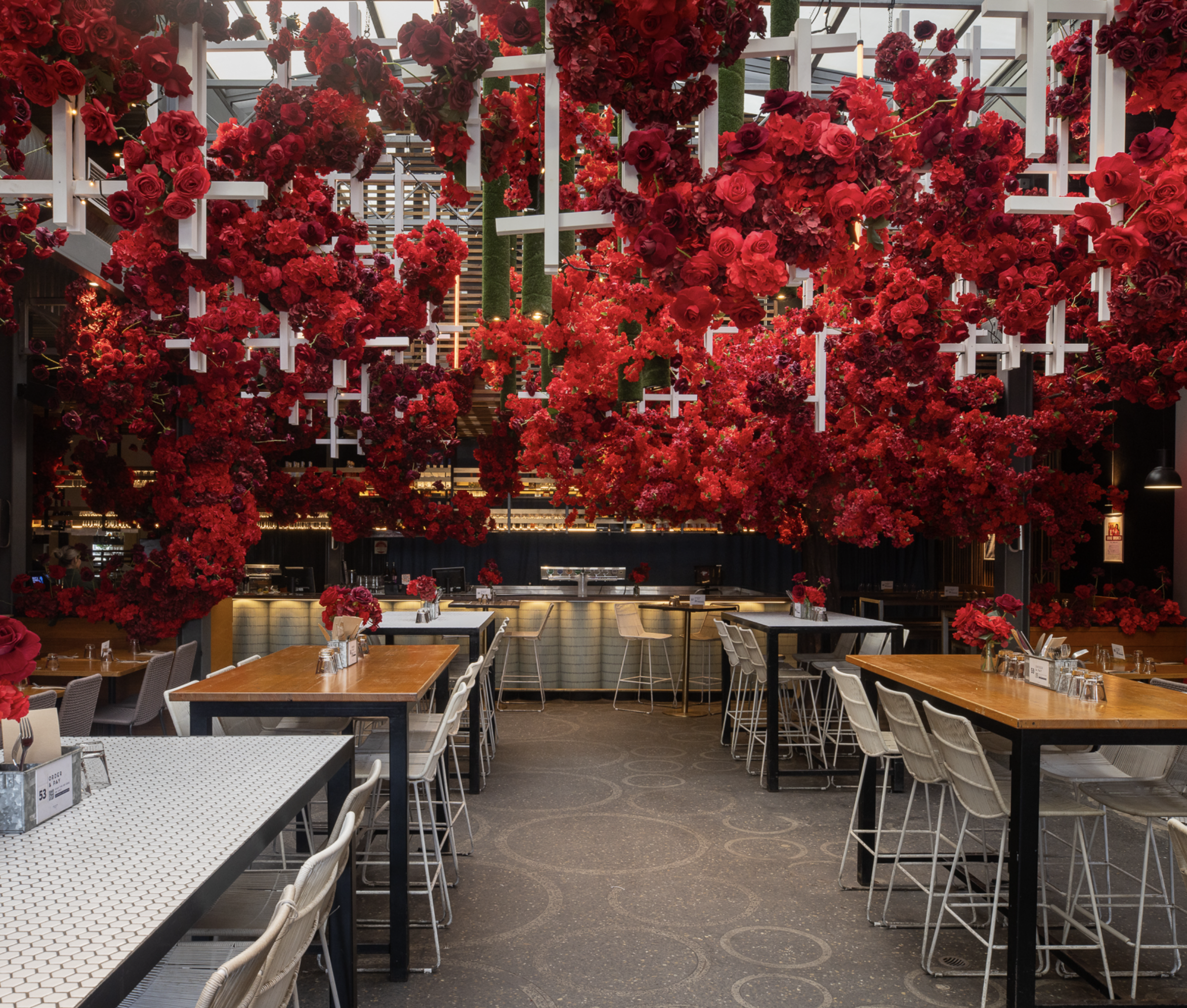 An indoor restaurant with a ceiling decorated with an abundant arrangement of red and pink flowers, including roses. The dining area has various tables and chairs, some with flowers in vases, and a bar or service counter in the background.