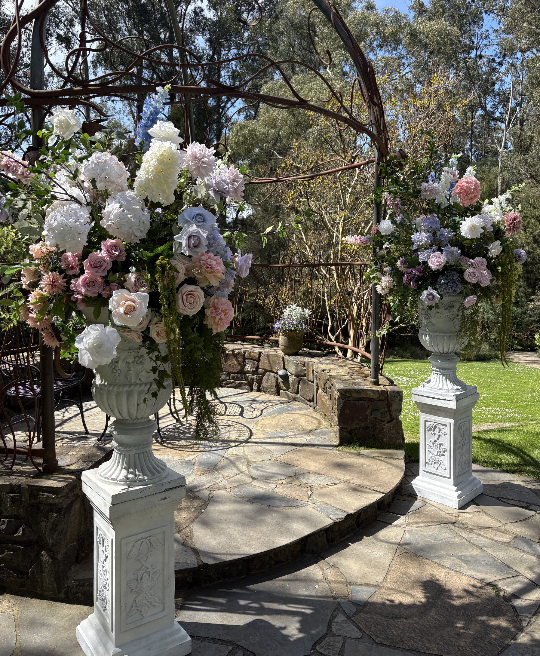 Beautiful outdoor garden scene with two large white classical vases filled with pastel-colored flowers on stone pedestals, set on a stone patio with a decorative metal garden arch and trees in the background.