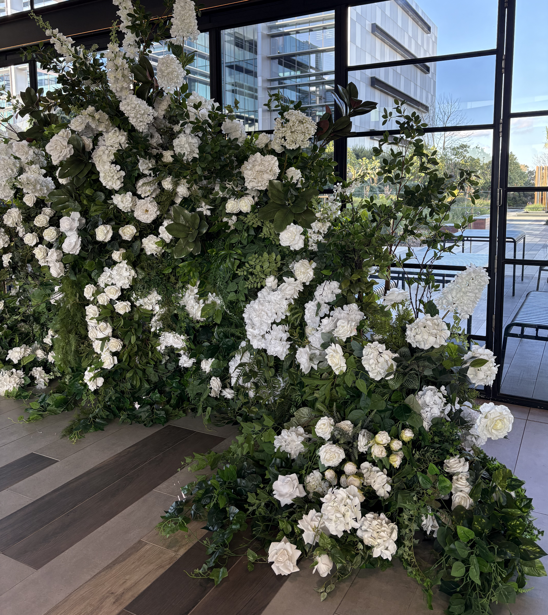 Large arrangement of white flowers with green leaves inside a room with glass walls and a view of modern buildings outside.