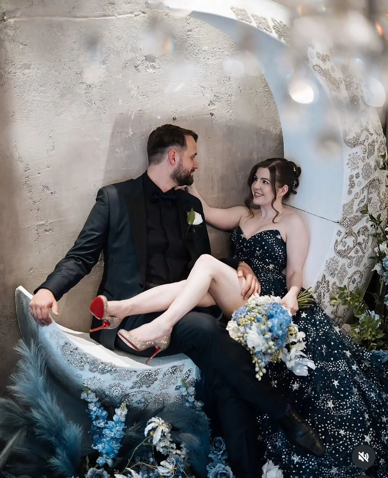 A bride and groom sitting on a decorative bench, with the bride sitting on the groom's lap and smiling at him. The bride is holding a bouquet of flowers, and both are dressed in formal attire. The setting is decorated with flowers and ornate fabrics.