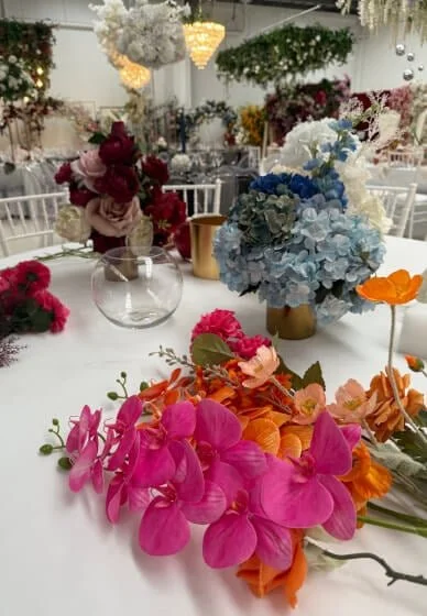 Colorful floral centerpieces with pink orchids, orange flowers, and blue and white hydrangeas on decorated banquet tables at a wedding or event venue.