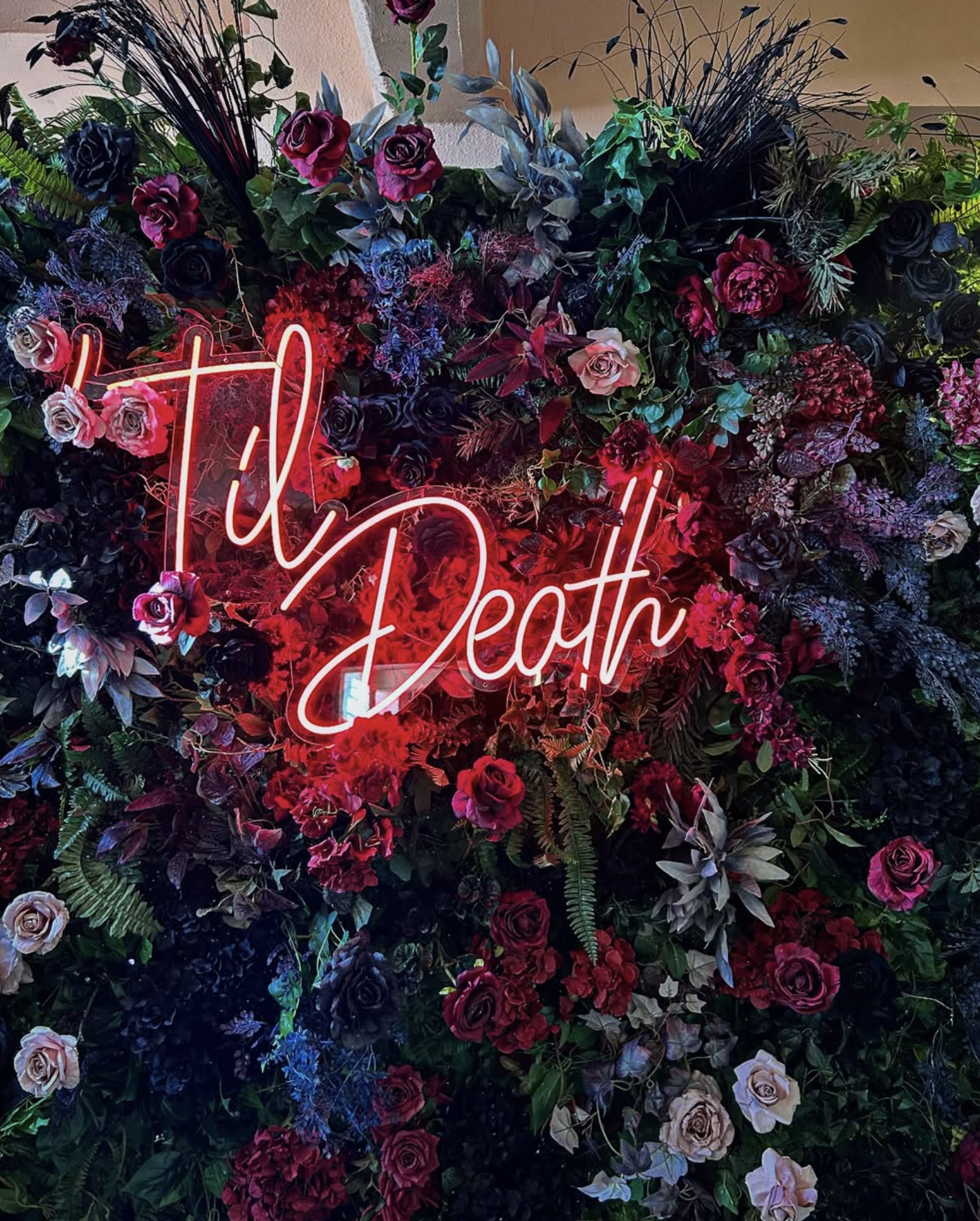 A floral arrangement with various roses and greenery, illuminated by a red neon sign that reads 'Till Death'.