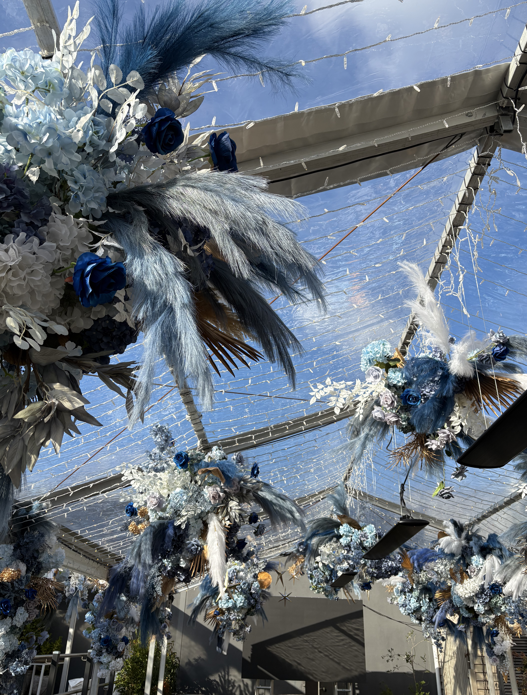 Decorative hanging floral arrangements with feathers and artificial flowers suspended from a transparent roof with string lights.