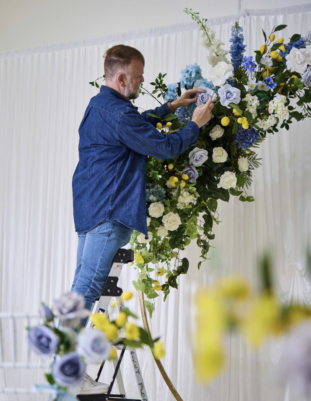 A man in a blue denim shirt arranging blue, white, and yellow flowers on a large floral display with a white backdrop.