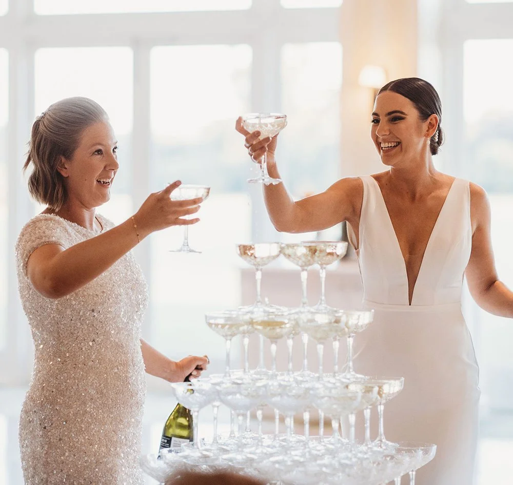 Two women celebrating with champagne glasses, toasting near a champagne tower in a bright room.