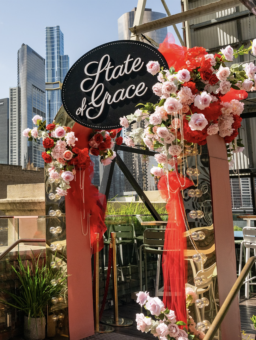 Outdoor event entrance decorated with pink and red flowers, red tulle, and pearl strings, with a sign that reads 'State of Grace' in cursive, set against a city skyline with tall modern buildings.