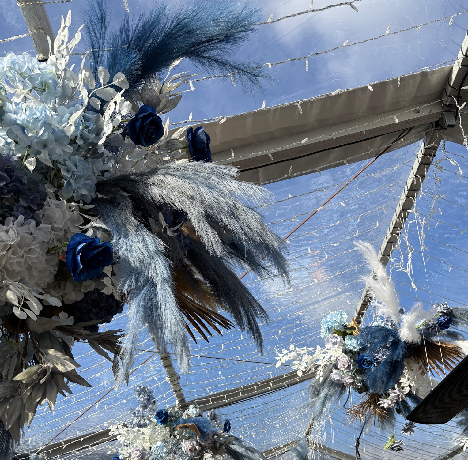 Decorative floral arrangements and feathers hanging from the ceiling under a transparent roof with string lights.