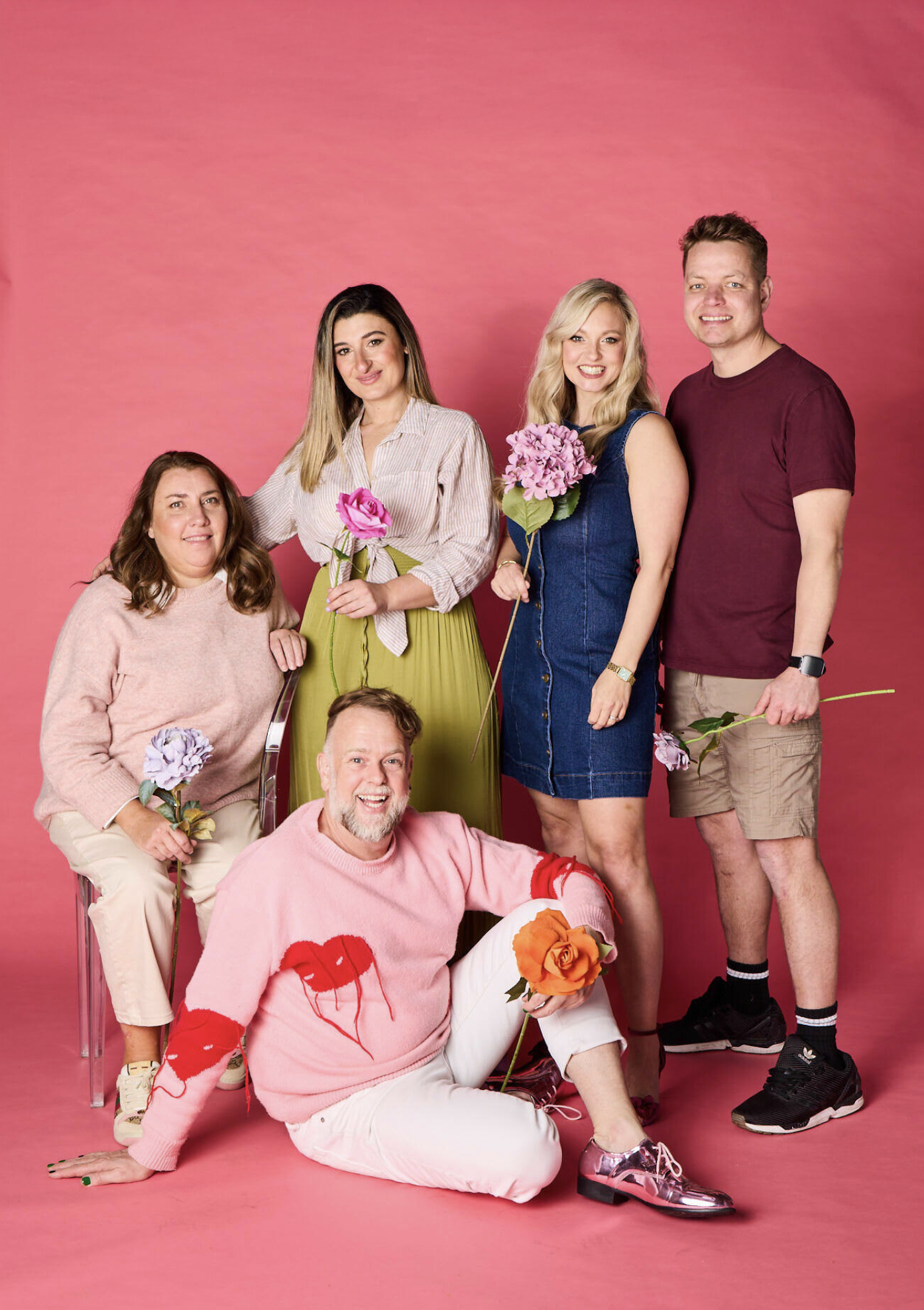 Group of six people posing against a pink background, holding colorful flowers.