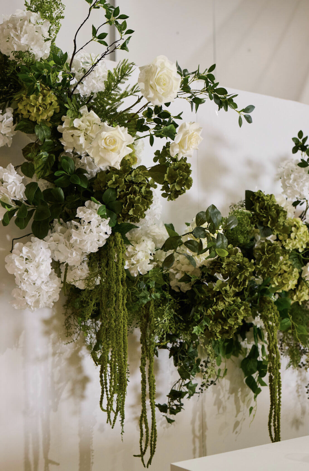 A floral arrangement with white roses, white hydrangeas, green leaves, and hanging green amaranthus on a white background.