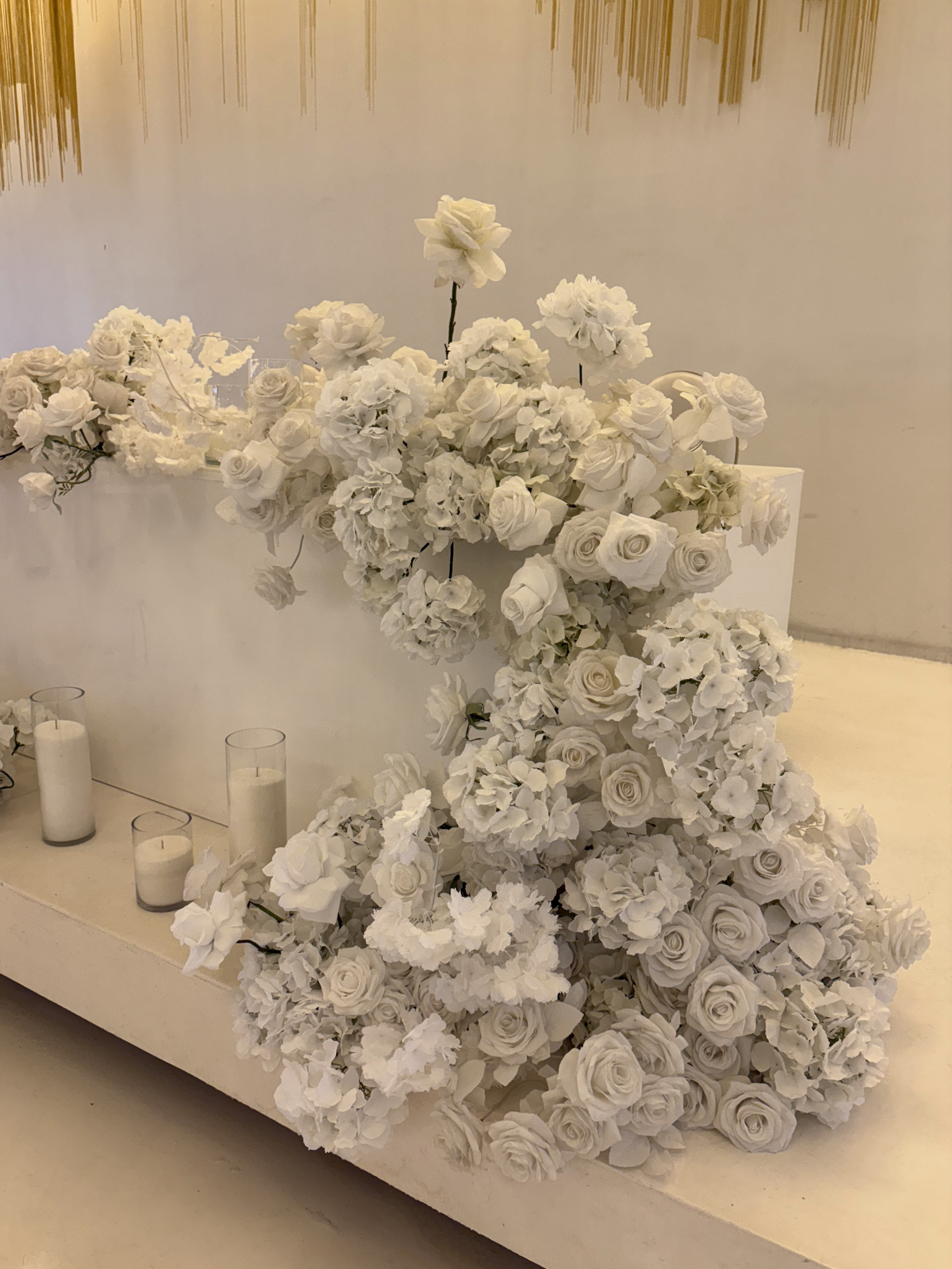 White floral arrangement with roses and hydrangeas on a white surface, with candles in glass holders nearby.