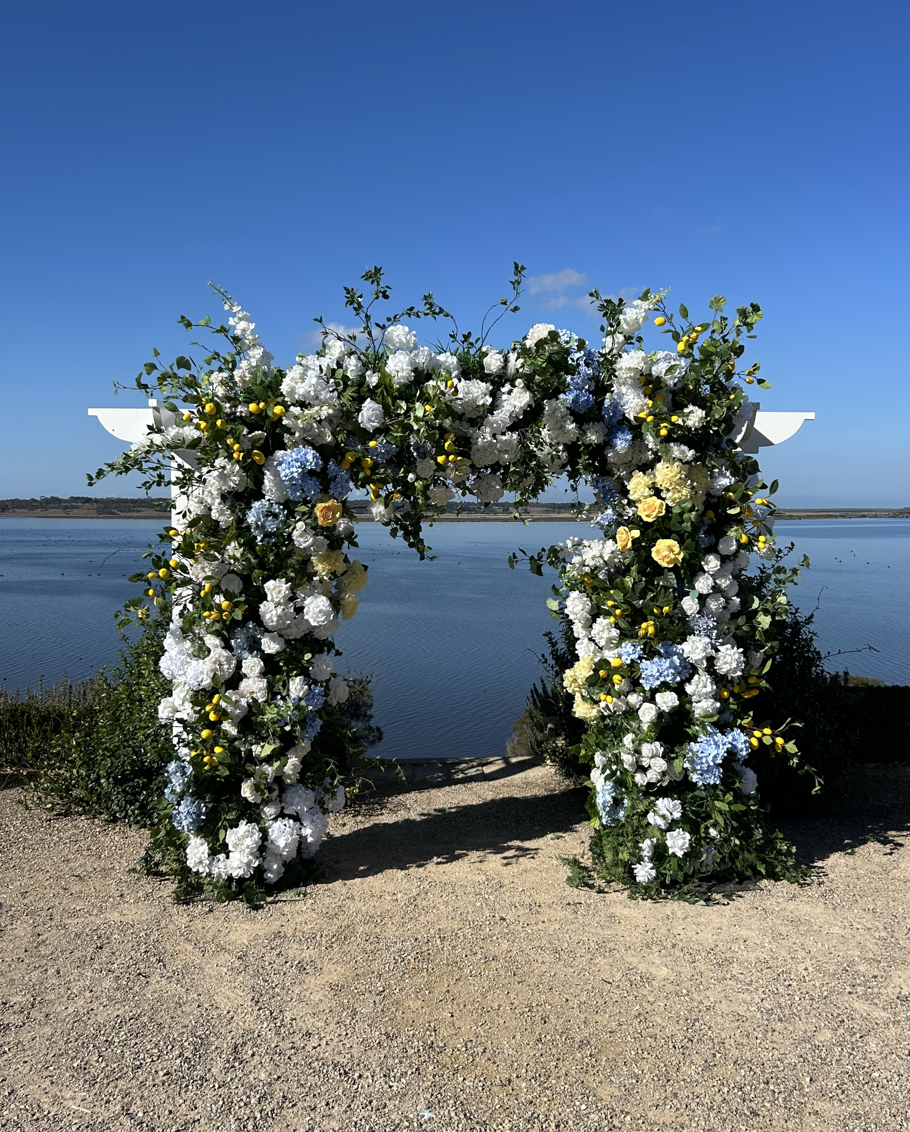 A floral arch decorated with white, yellow, and light blue flowers, set against a lakeside with a clear blue sky.