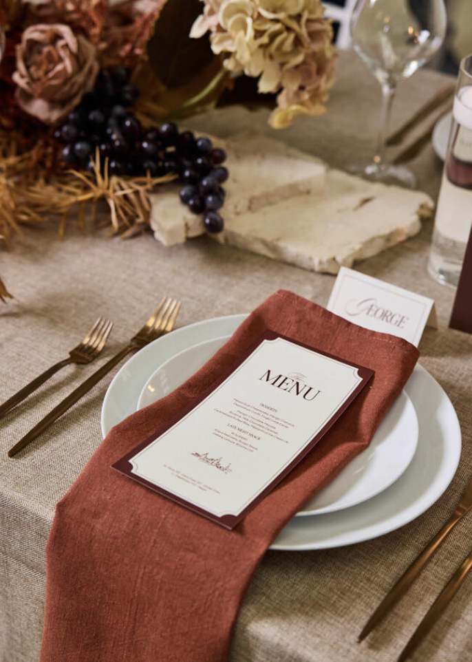 Elegant table setting with a white plate, a menu card on a red napkin, gold cutlery, and a floral centerpiece with dried flowers, grapes, and a marble slab in the background.