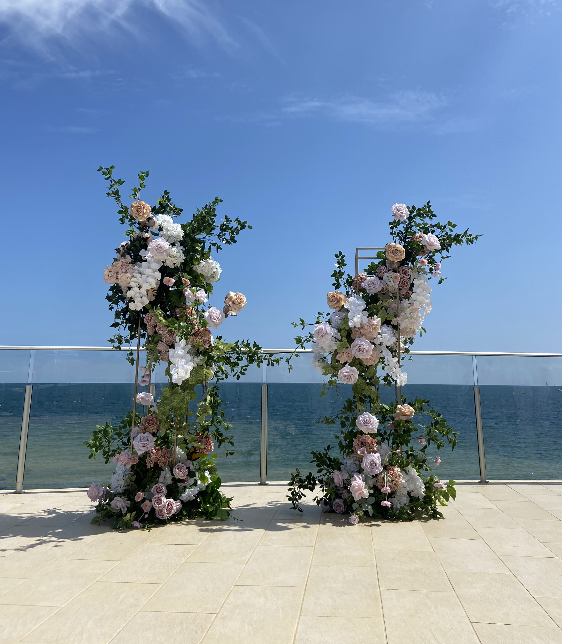 Floral wedding arch with pastel pink, white, and cream roses, and green foliage, set against the ocean and blue sky.