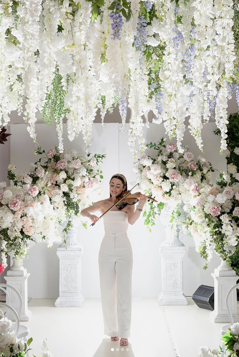 A woman in a white dress playing a violin amid an elaborate floral arrangement with white and pink flowers and hanging white and purple flowers.