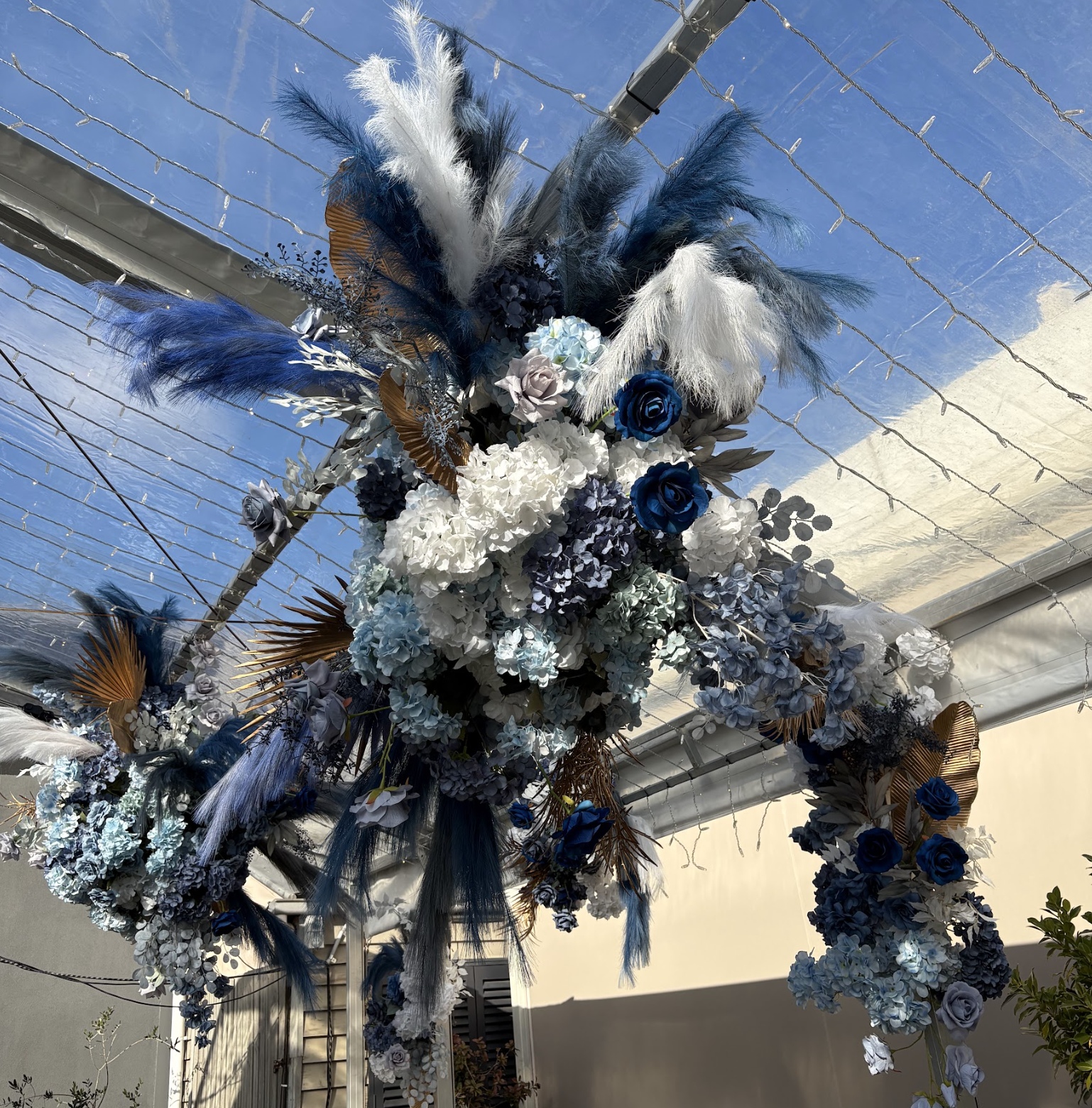 A floral decoration with white, blue, and gray flowers, accented with blue and white feathers, arranged in an arch under a transparent roof.