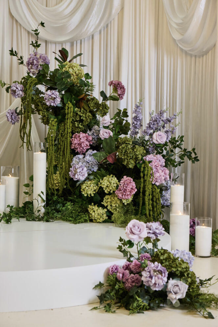 A floral arrangement with pink, purple, and green flowers, candles, and greenery on a white surface, set against a cream-colored draped curtain background.