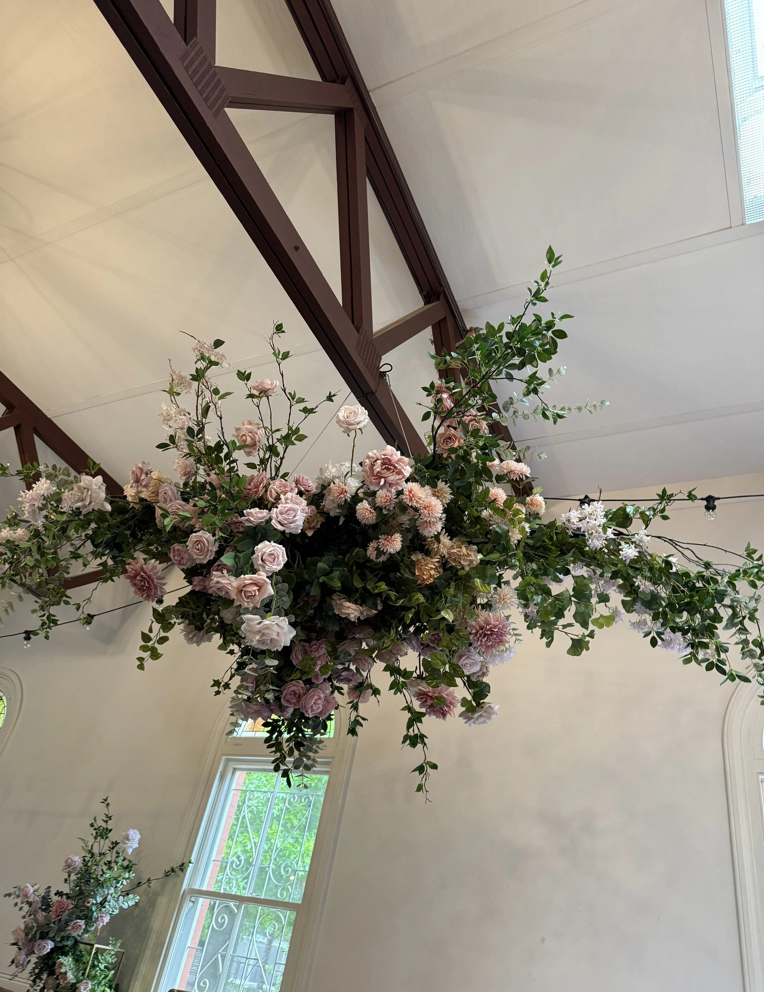 A floral arrangement with pink and white roses and greenery hanging from the ceiling of a room with high ceilings and windows.