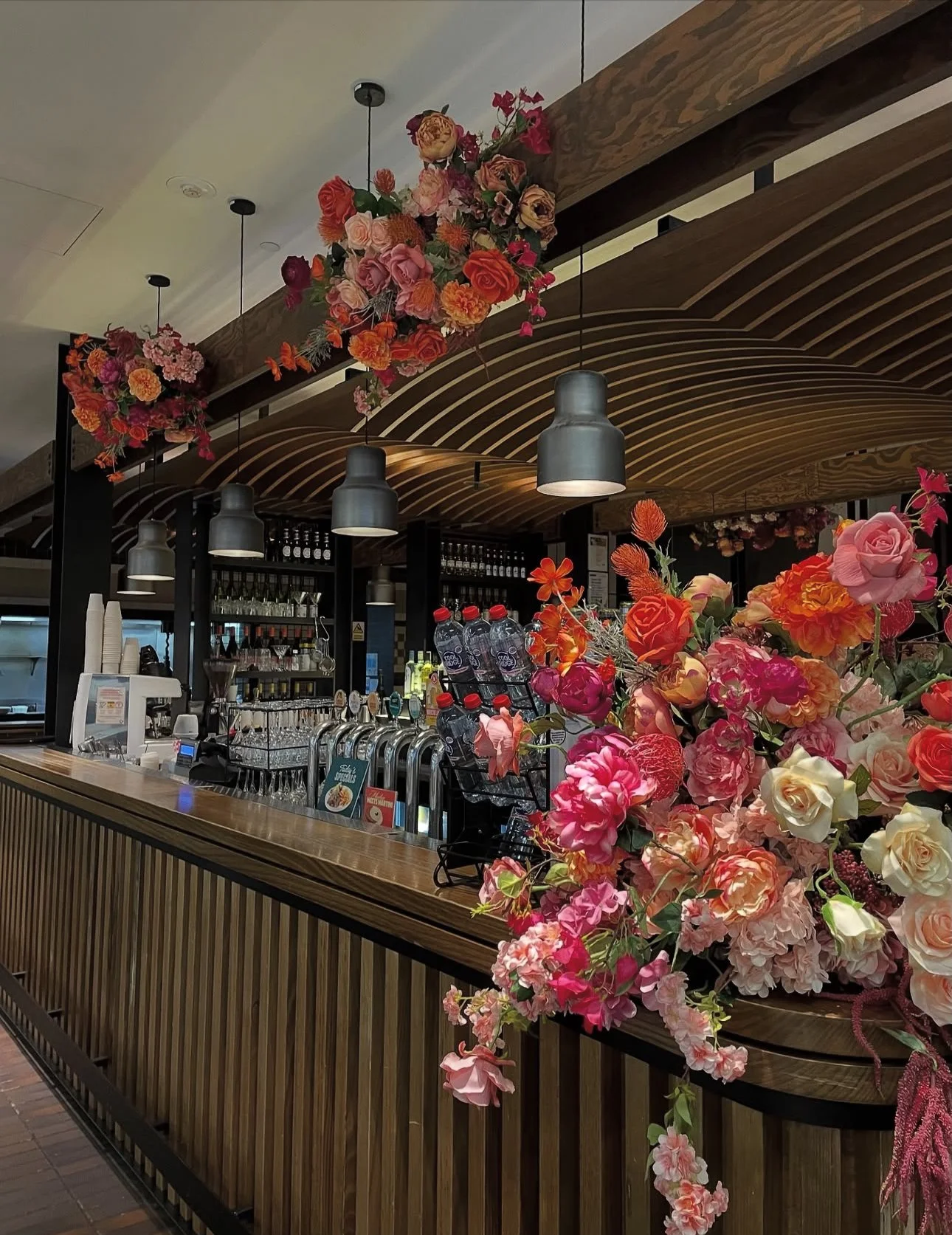 Inside a coffee shop or cafe with a wooden counter, black pendant lights, and floral decorations suspended from the ceiling and on the counter.