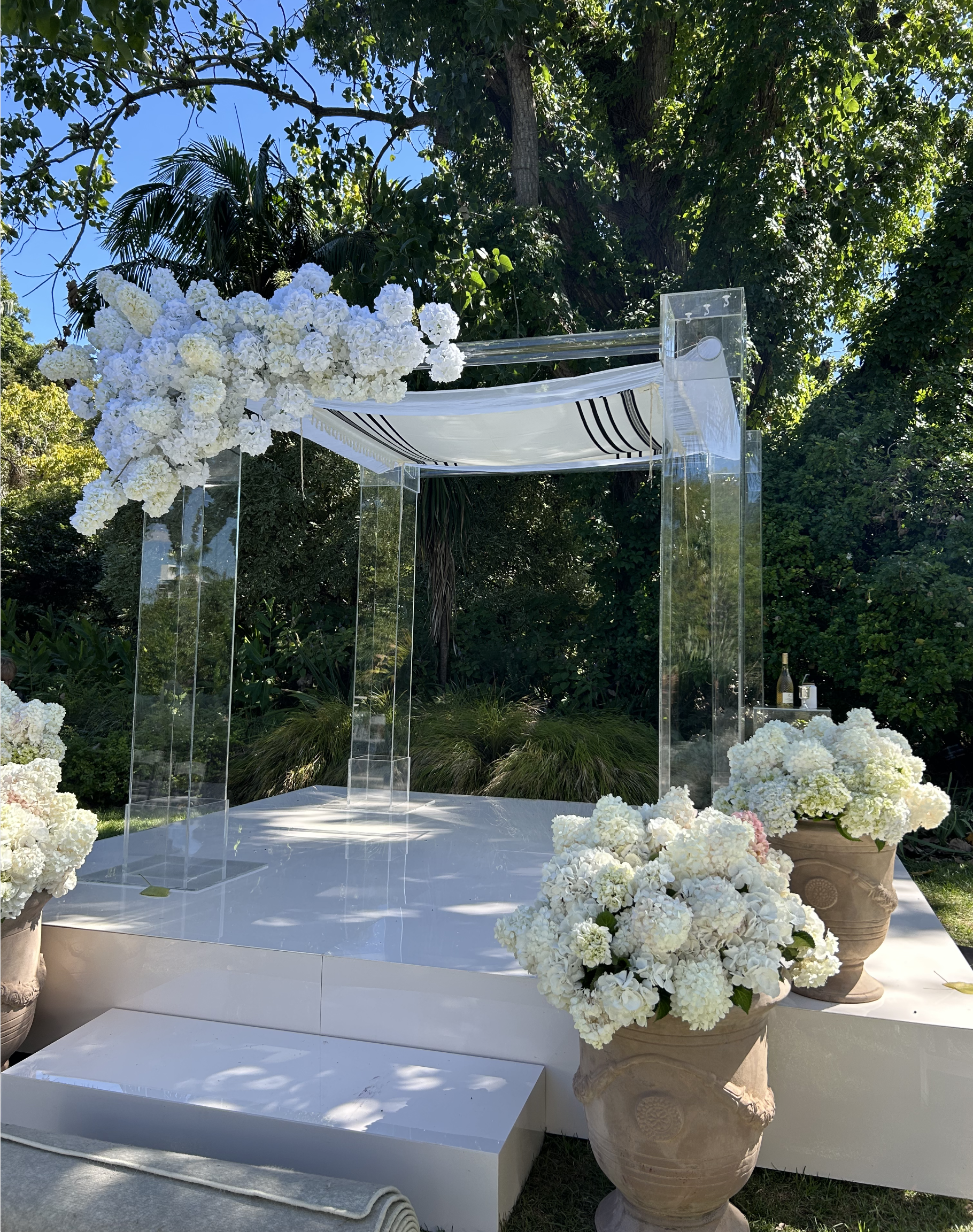 Wedding ceremony stage decorated with white flowers, clear acrylic pillars, and white and black drapery, set outdoors under trees.