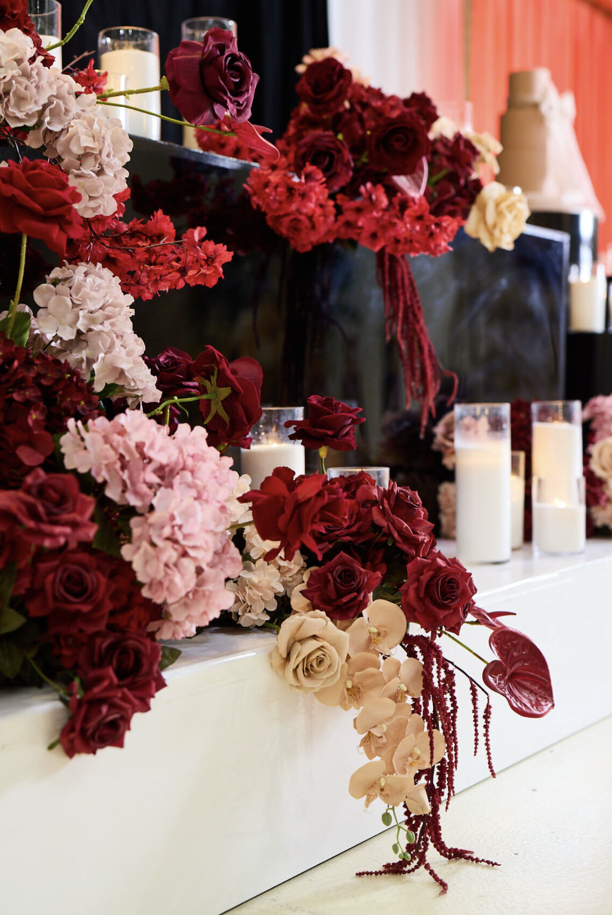 Arrangement of red, pink, and white roses, hydrangeas, orchids, and red anthuriums with candles, placed on a white surface, with a black piano and red curtains in the background.
