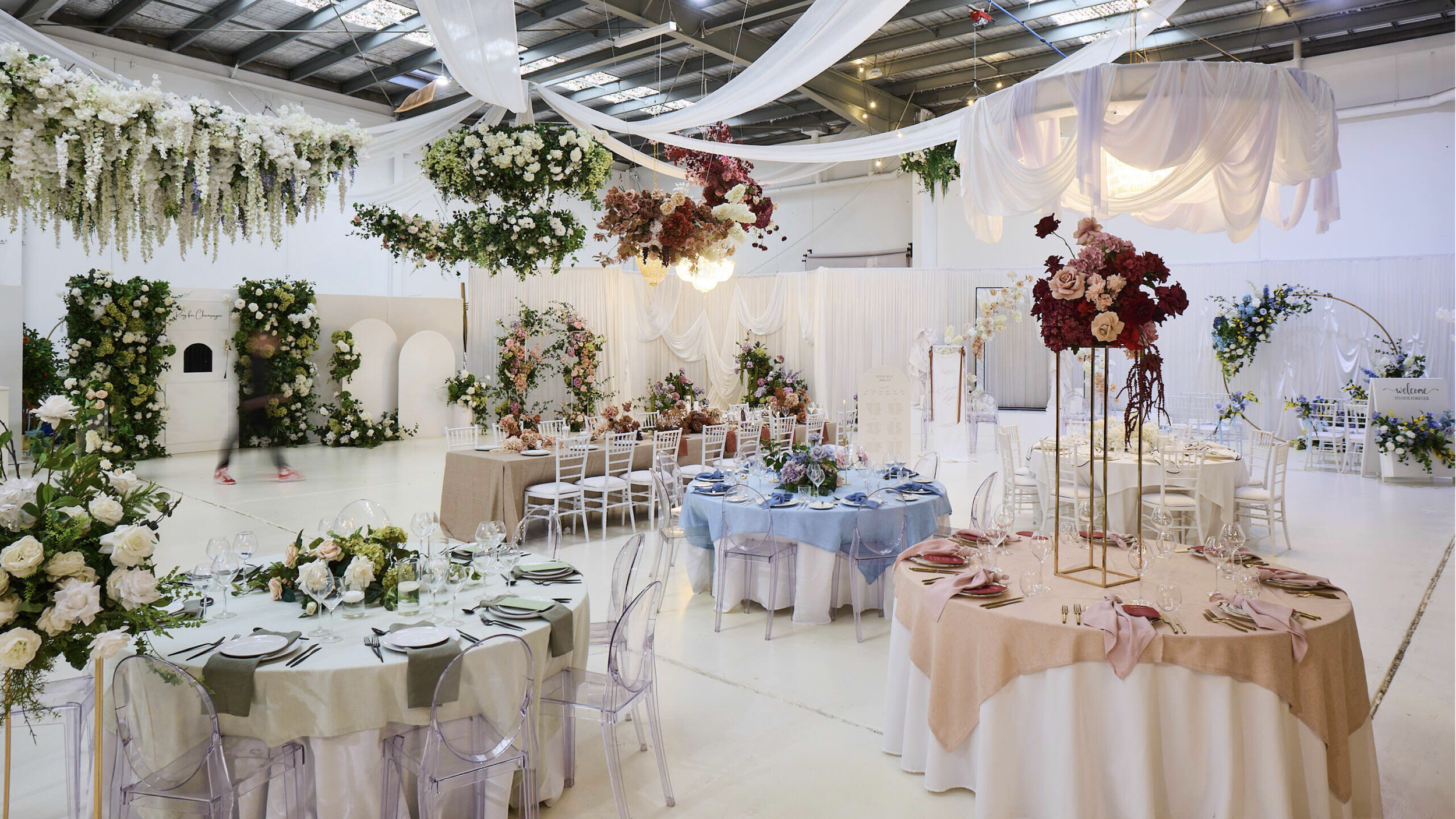 Elegant wedding reception with decorated tables, floral centerpieces, and white drapes in a spacious venue.