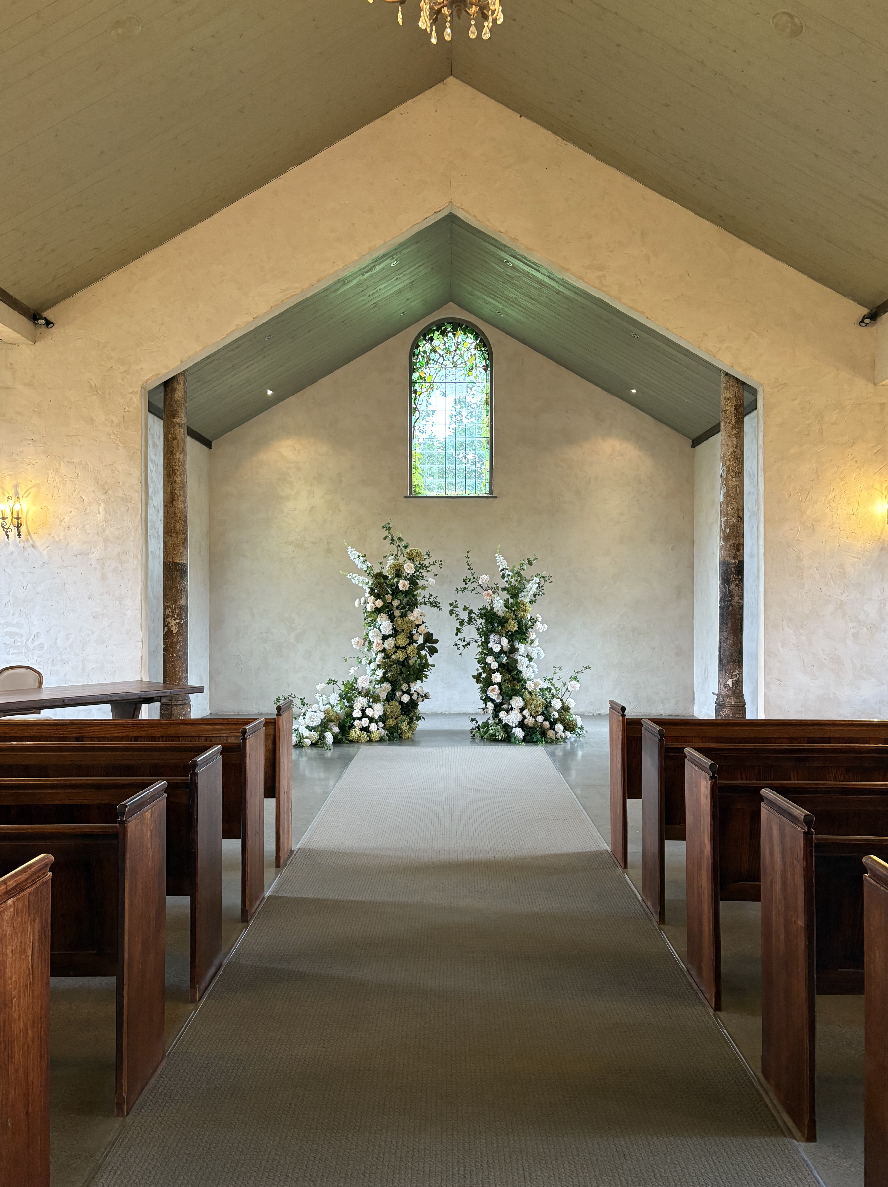 Interior of a small wedding chapel with wooden pews, floral arrangements, a stained glass window, and a chandelier.