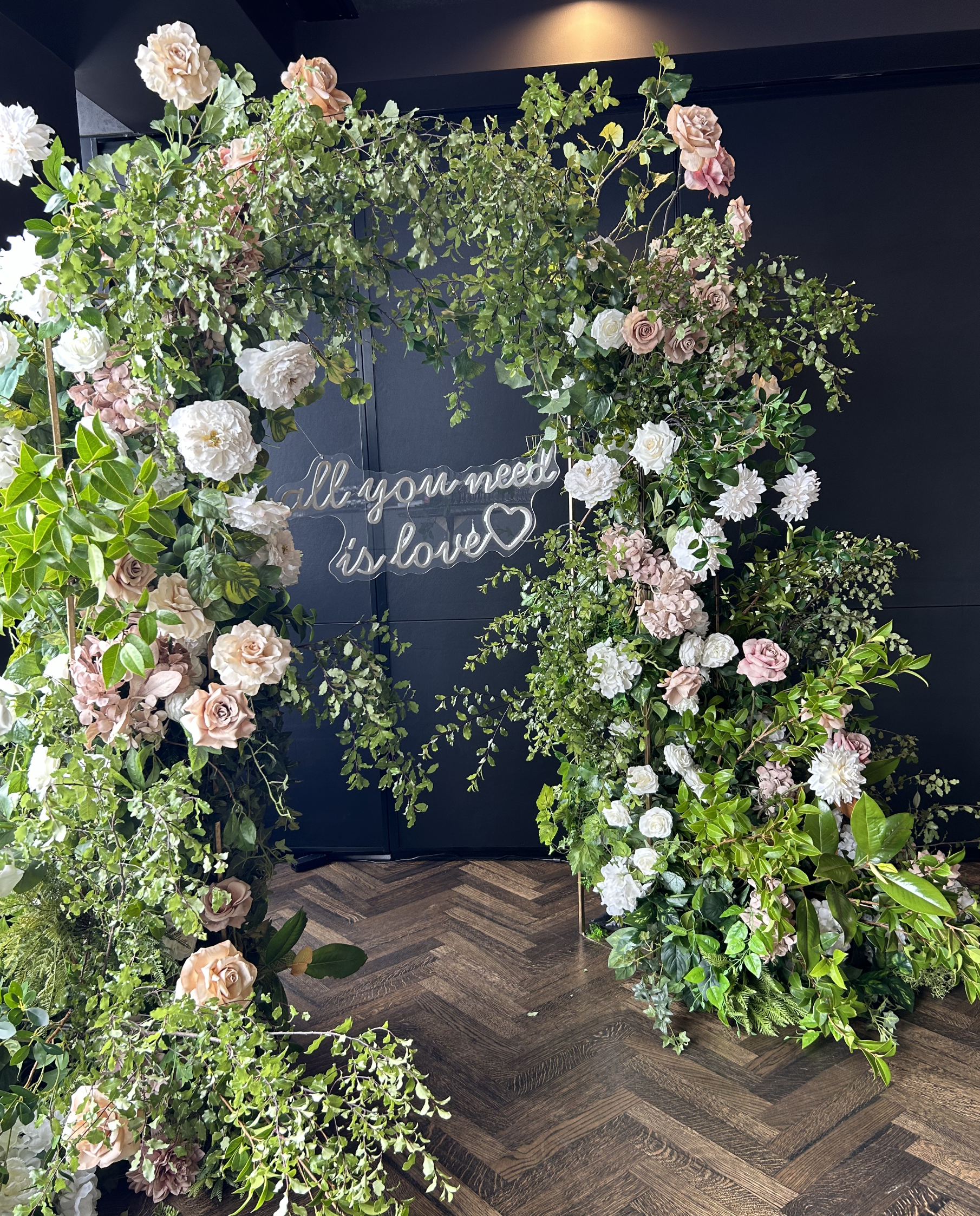 Floral arch with roses and greenery surrounding a sign that says 'all you need is love'